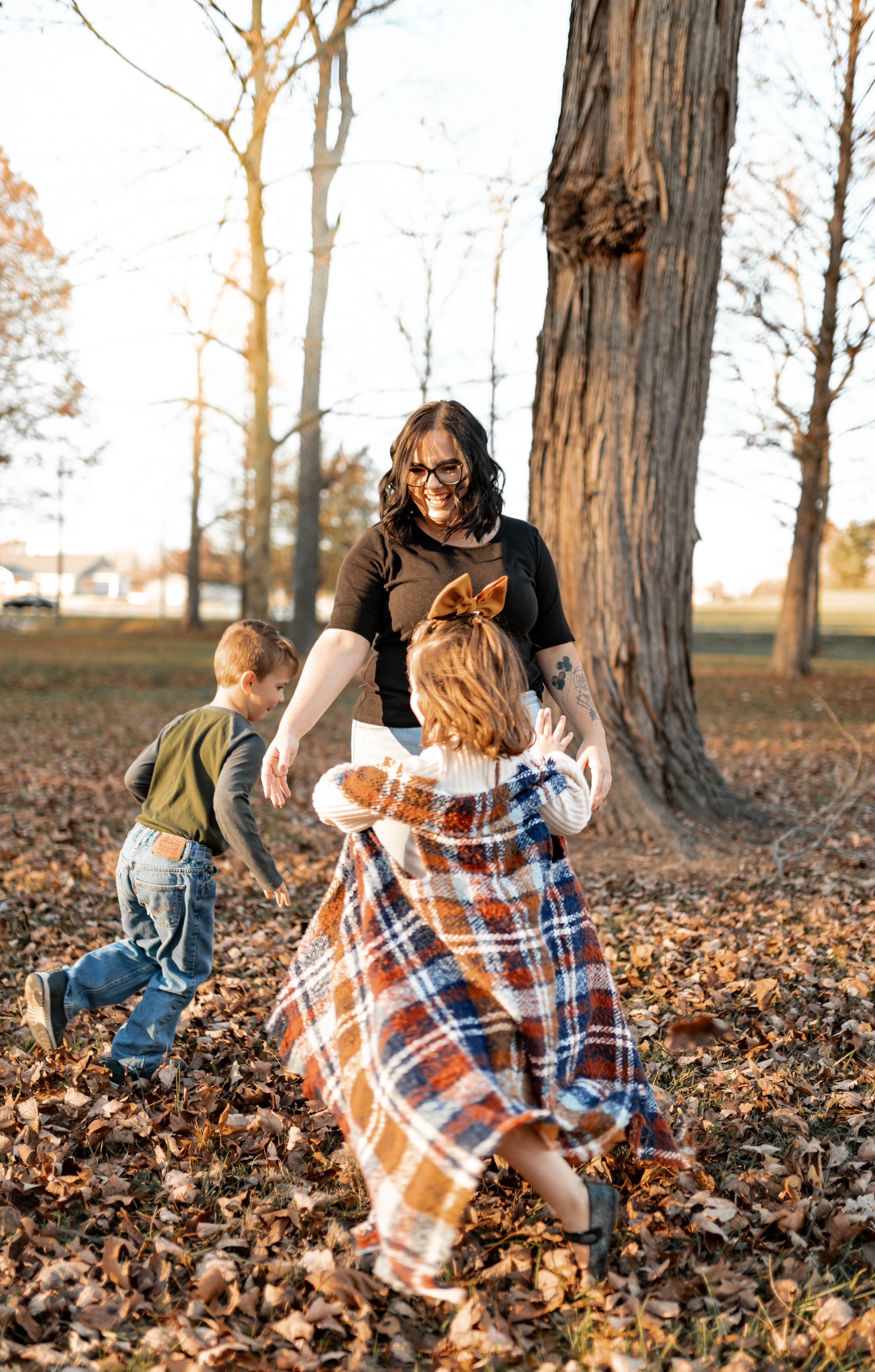 A woman playing with two children in a park during fall, surrounded by fallen leaves and tall trees.