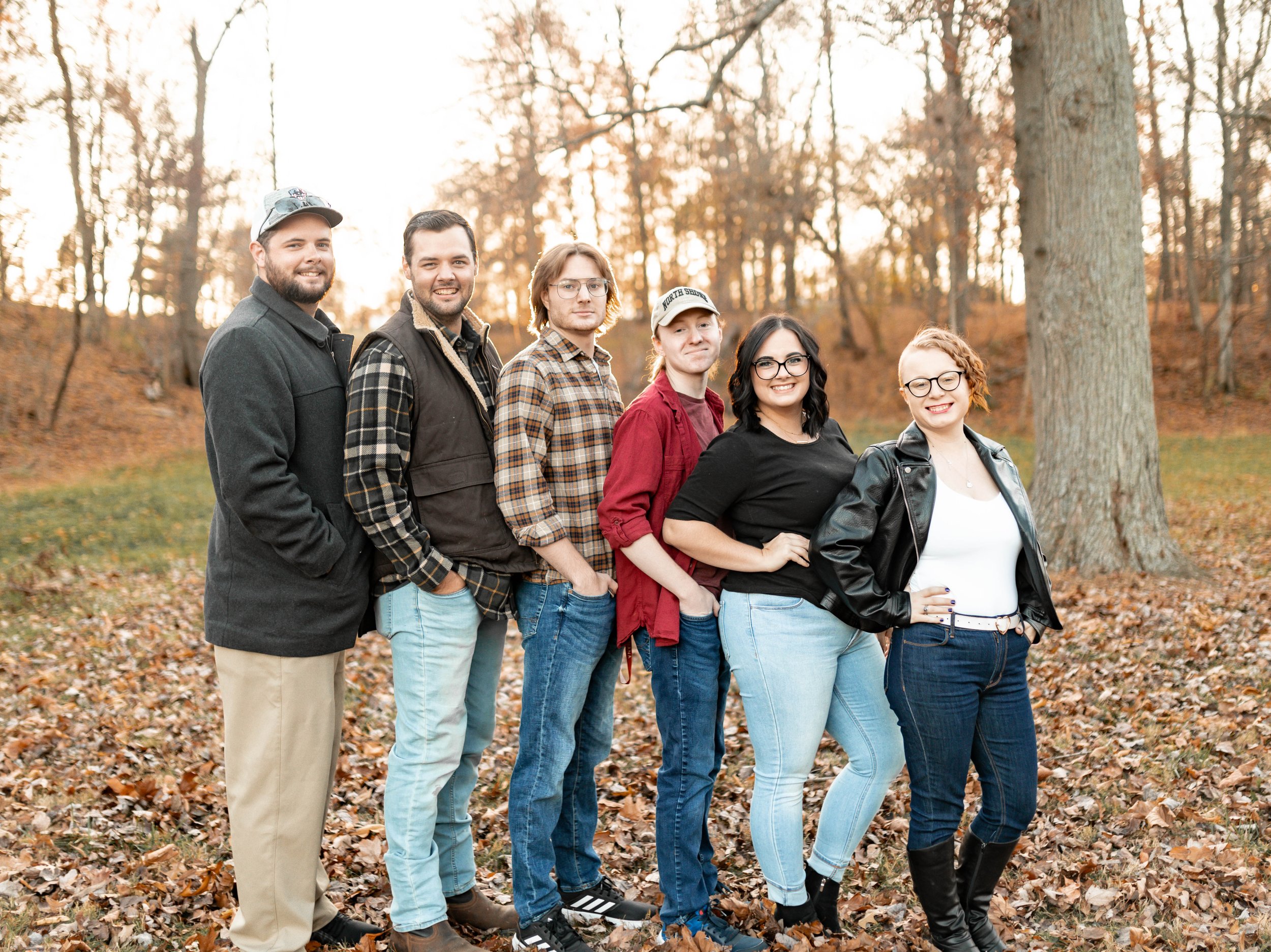 Group of six young adults standing outdoors in a wooded park with fallen leaves, smiling at the camera during autumn sunset.