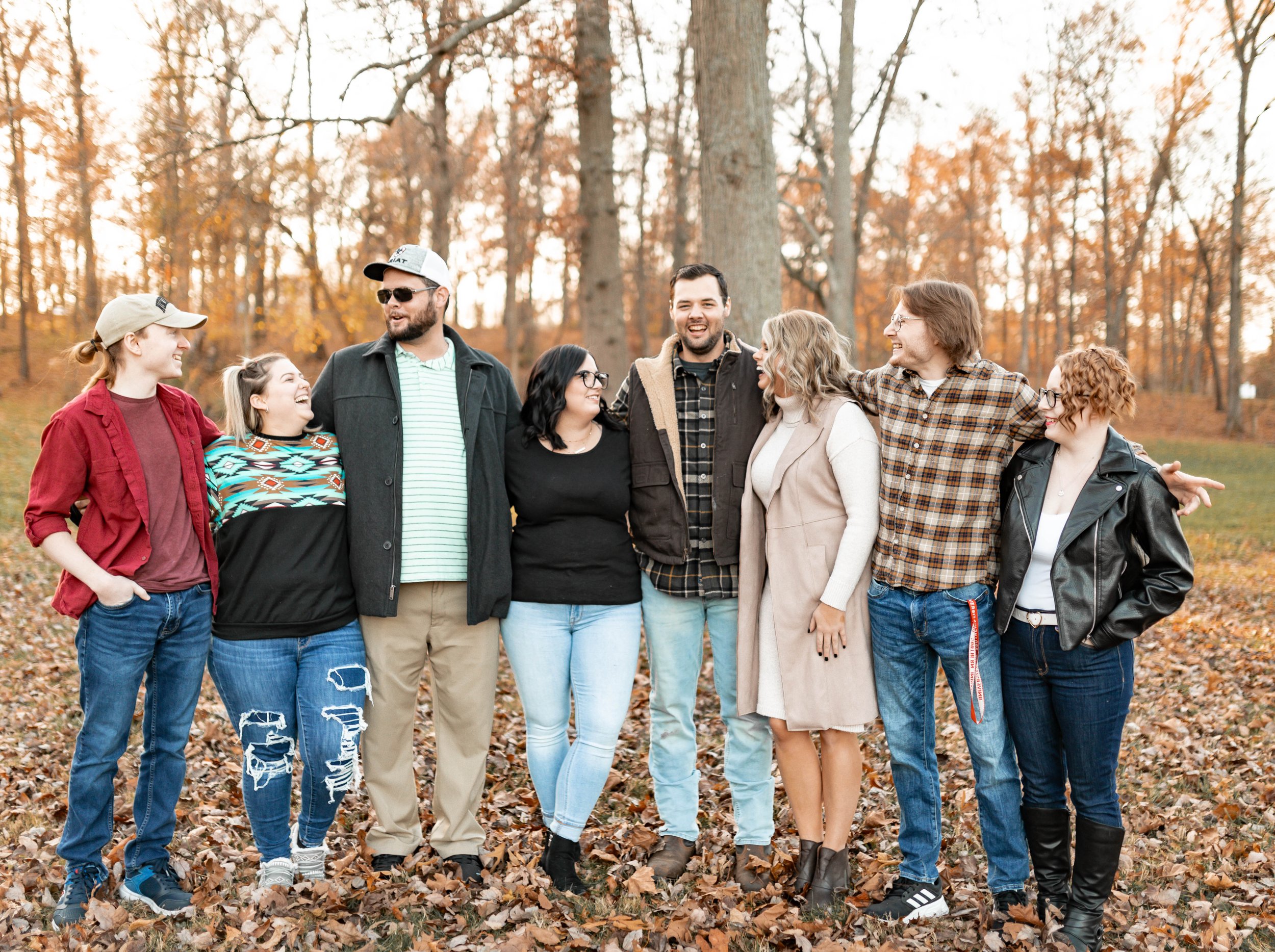 Group of eight friends standing outdoors on autumn leaves, smiling and enjoying each other's company.