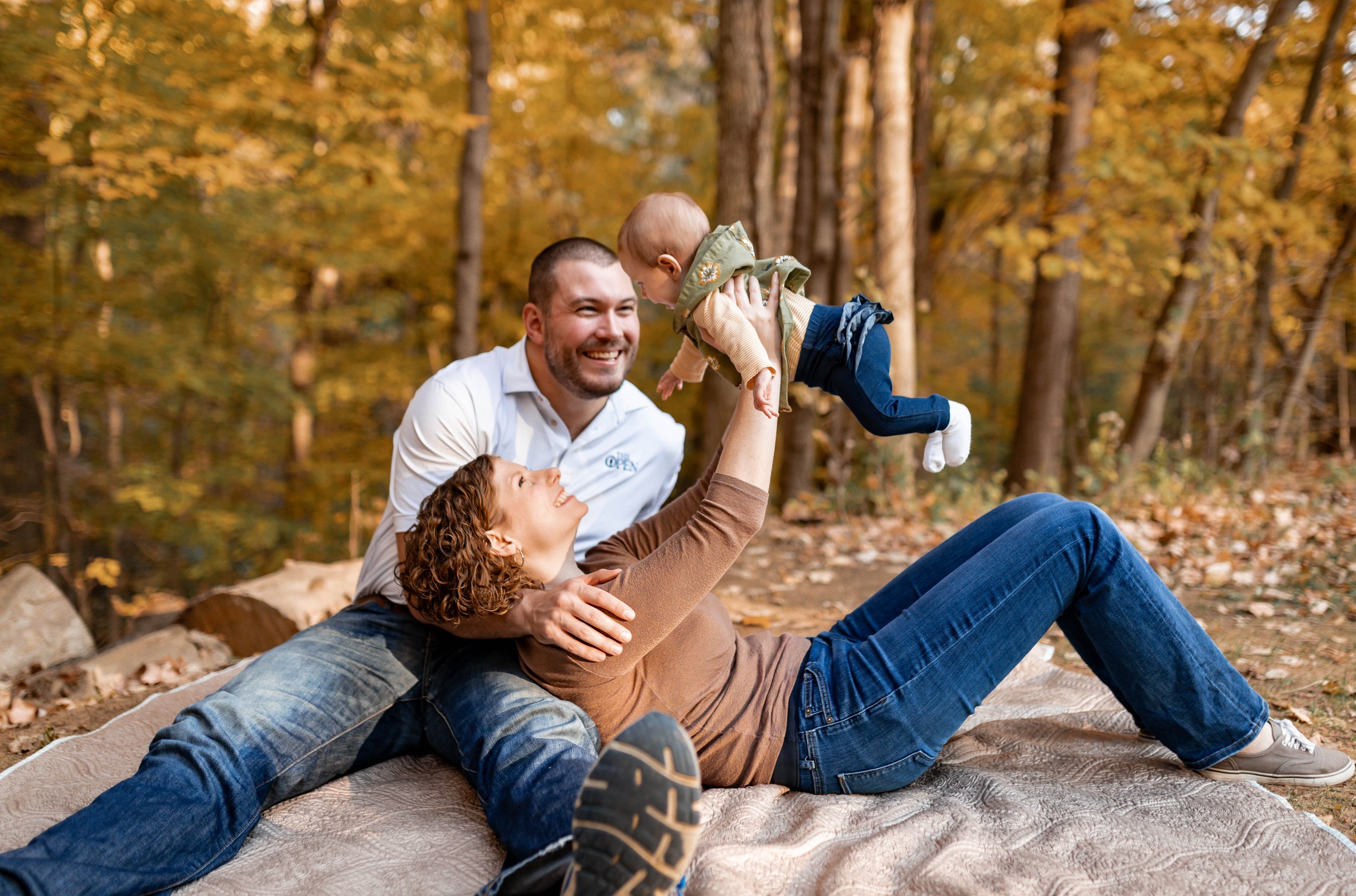 Family playing outdoors on a blanket in a forest during fall, with yellow and orange leaves, a woman lying on the ground lifting a baby and a man smiling in the background.