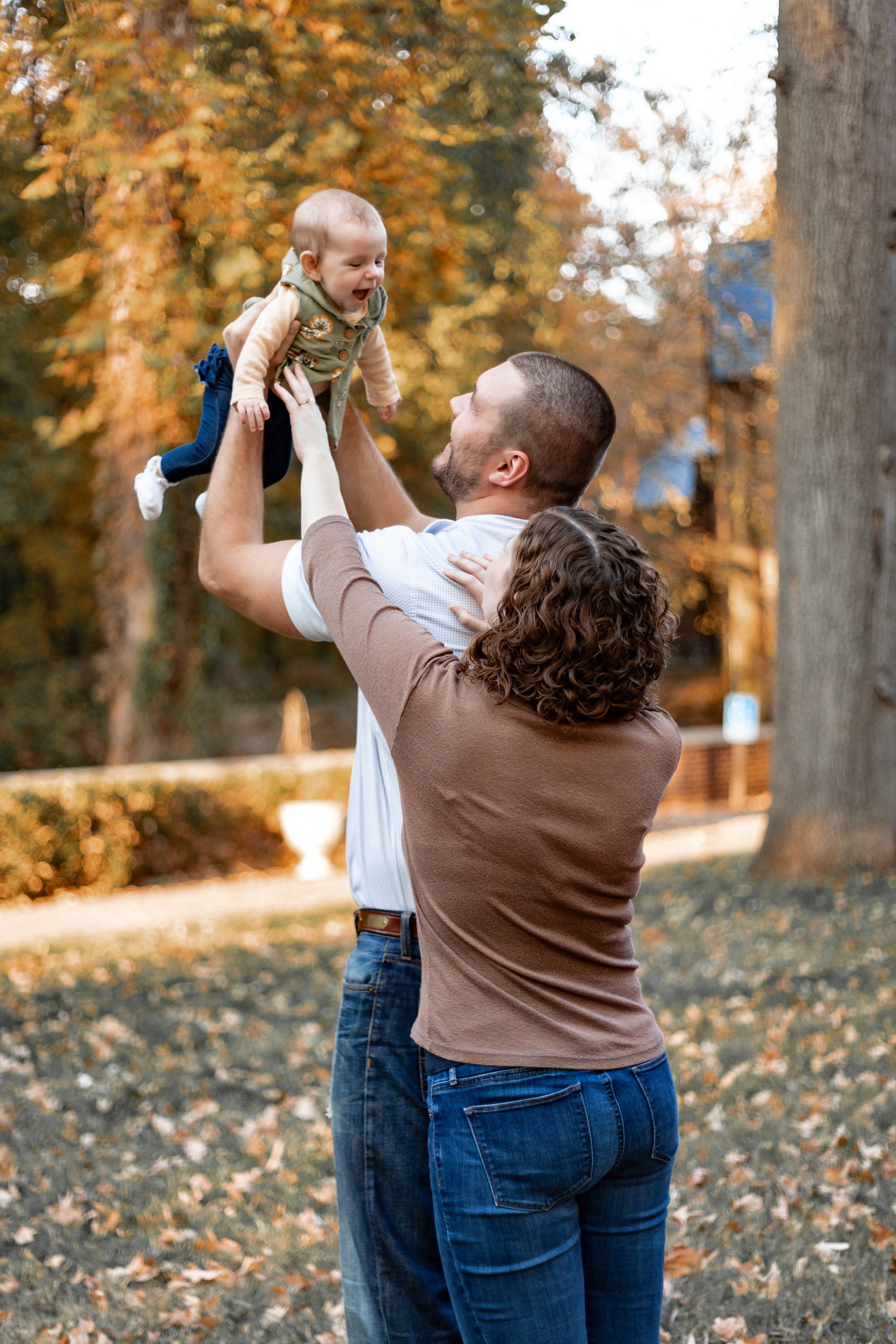 A man and woman playing with a smiling baby outdoors in a park during autumn.