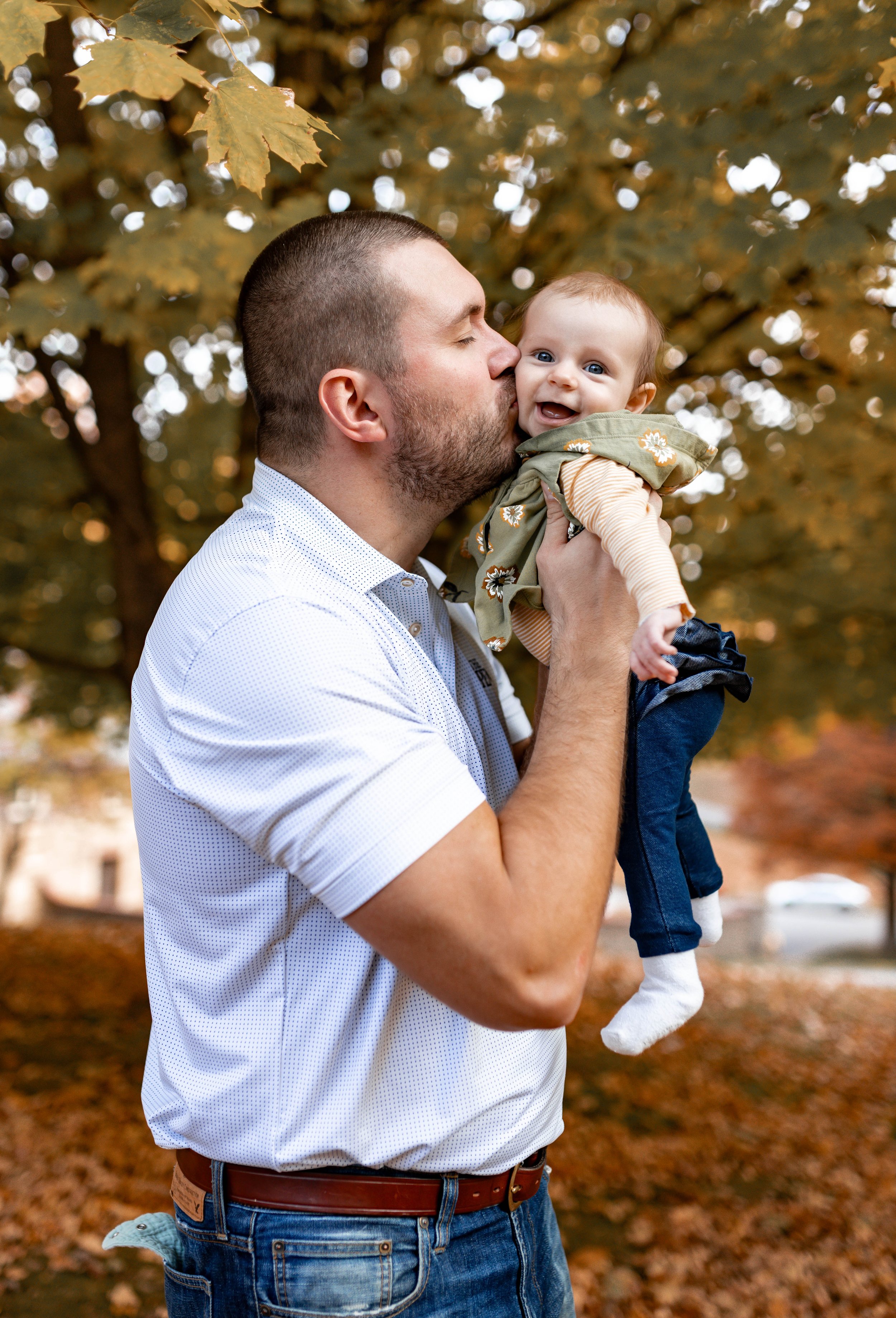 A man holding a smiling baby outdoors during autumn, surrounded by yellow and orange leaves.