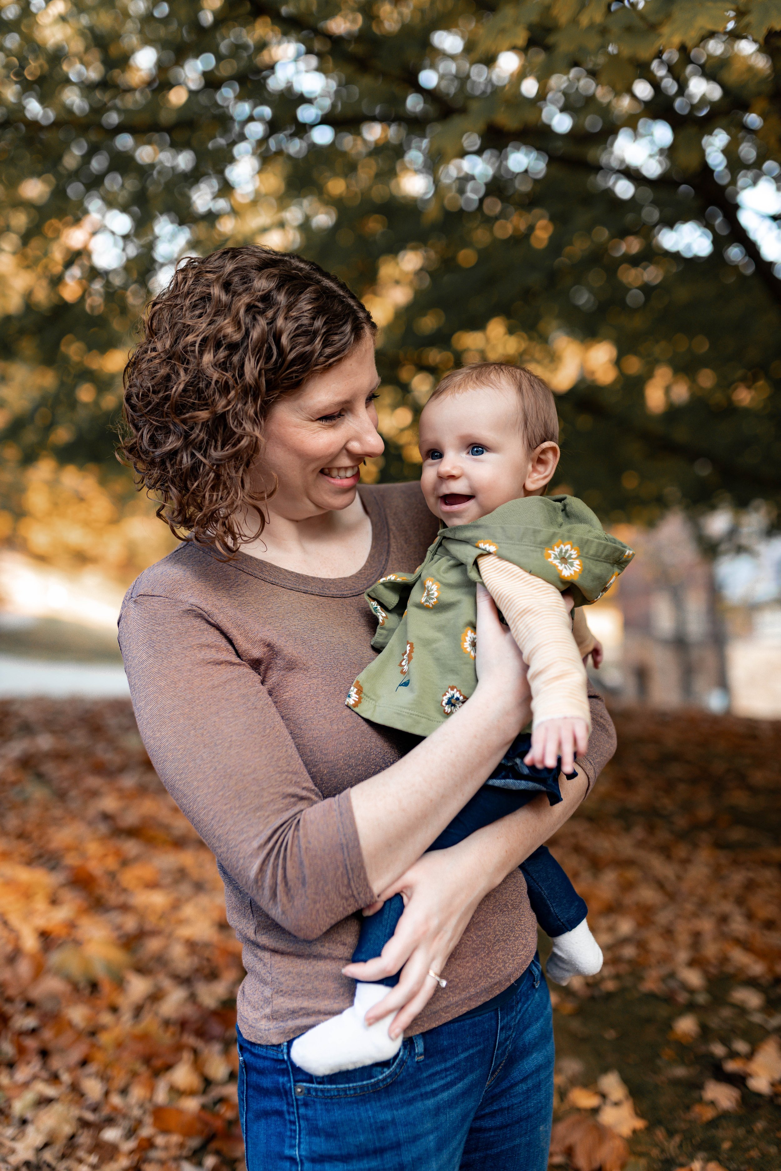 A woman with curly brown hair smiling and holding a baby outdoors during fall with orange and yellow leaves on the ground and trees in the background.