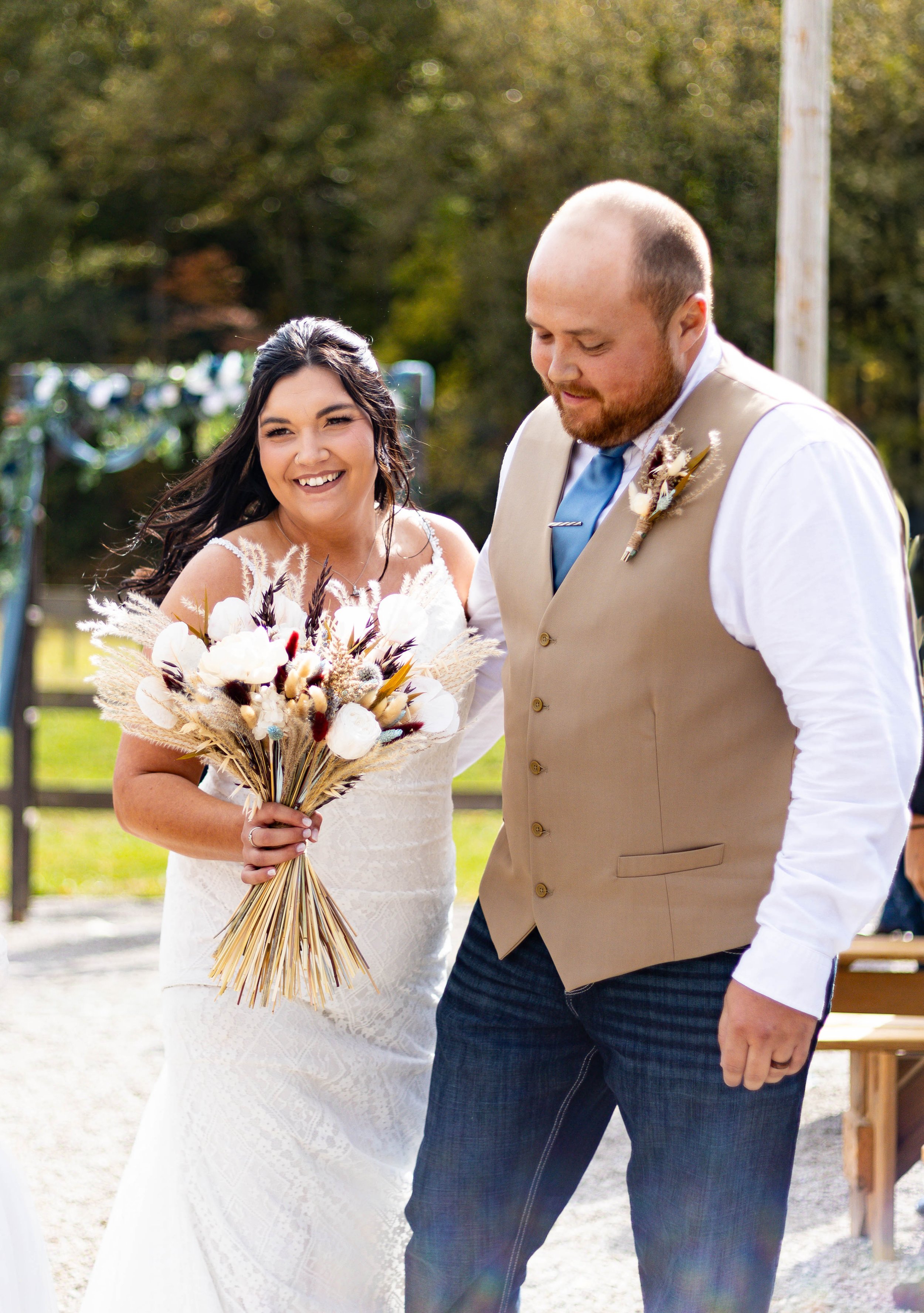 A bride with long dark hair and a white lace wedding dress holding a bouquet of dried flowers smiling at a groom with a beard, wearing a beige vest over a white shirt and a blue tie, during an outdoor wedding ceremony.