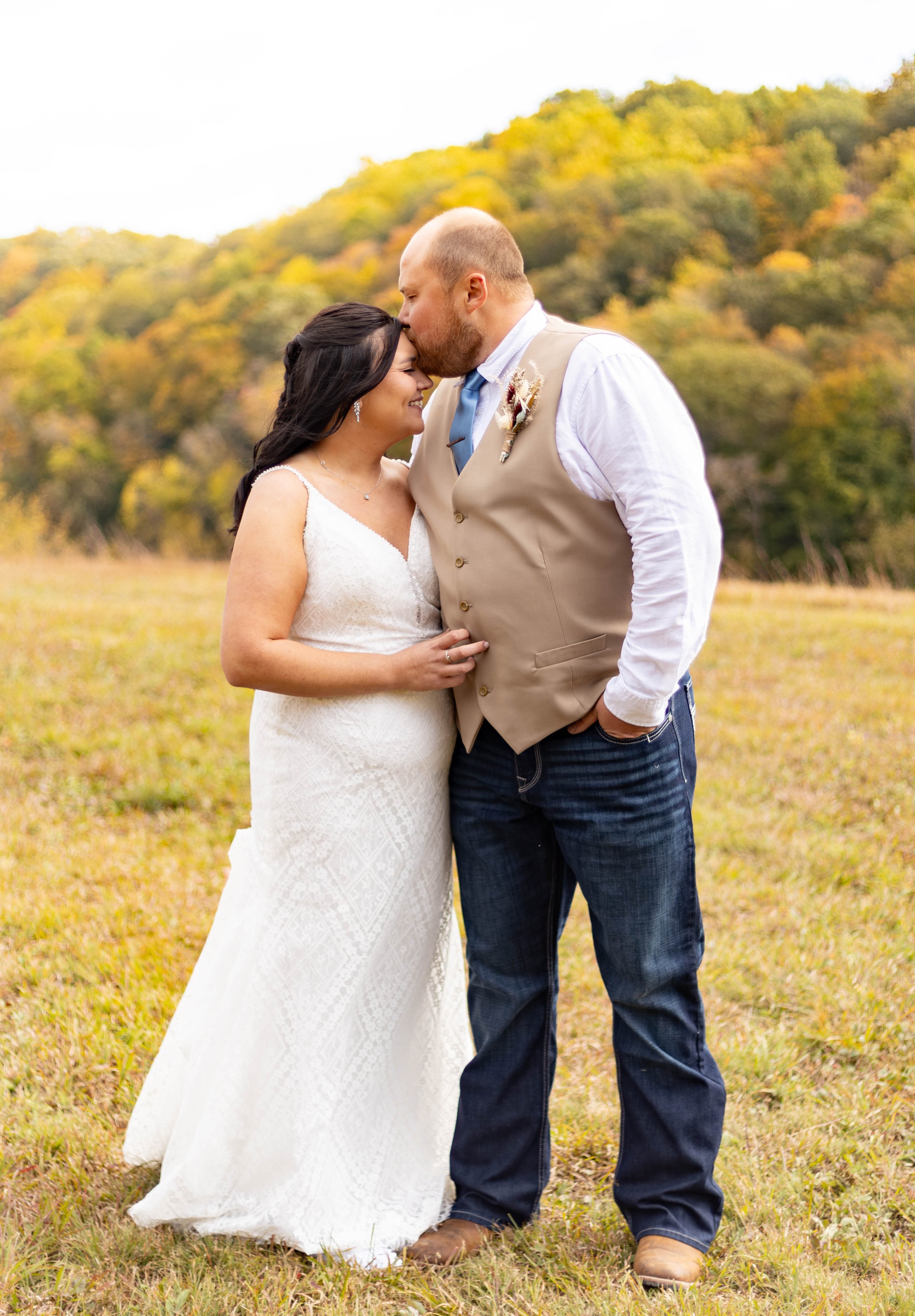 A bride and groom sharing a kiss outdoors during their wedding, with a backdrop of fall foliage in the distance.