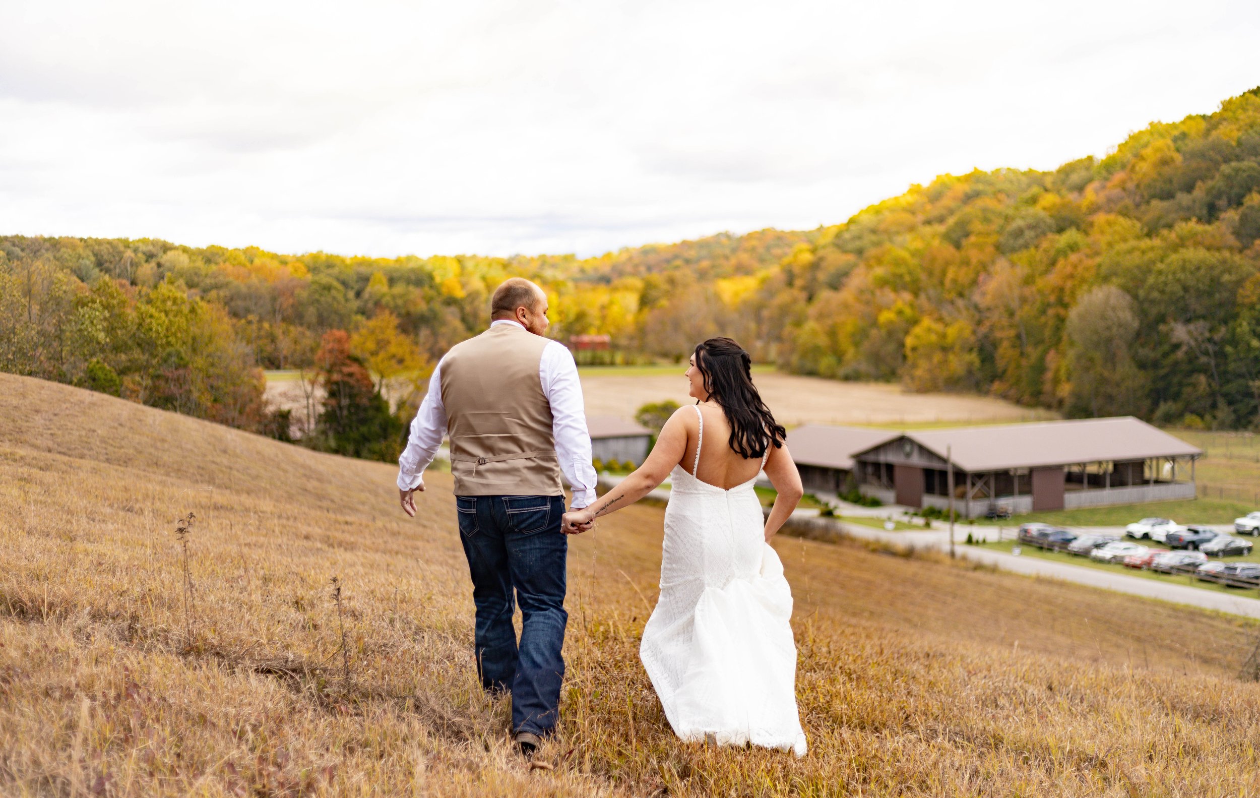 A couple holding hands walking on a grassy hillside with trees and a barn in the background, during autumn.