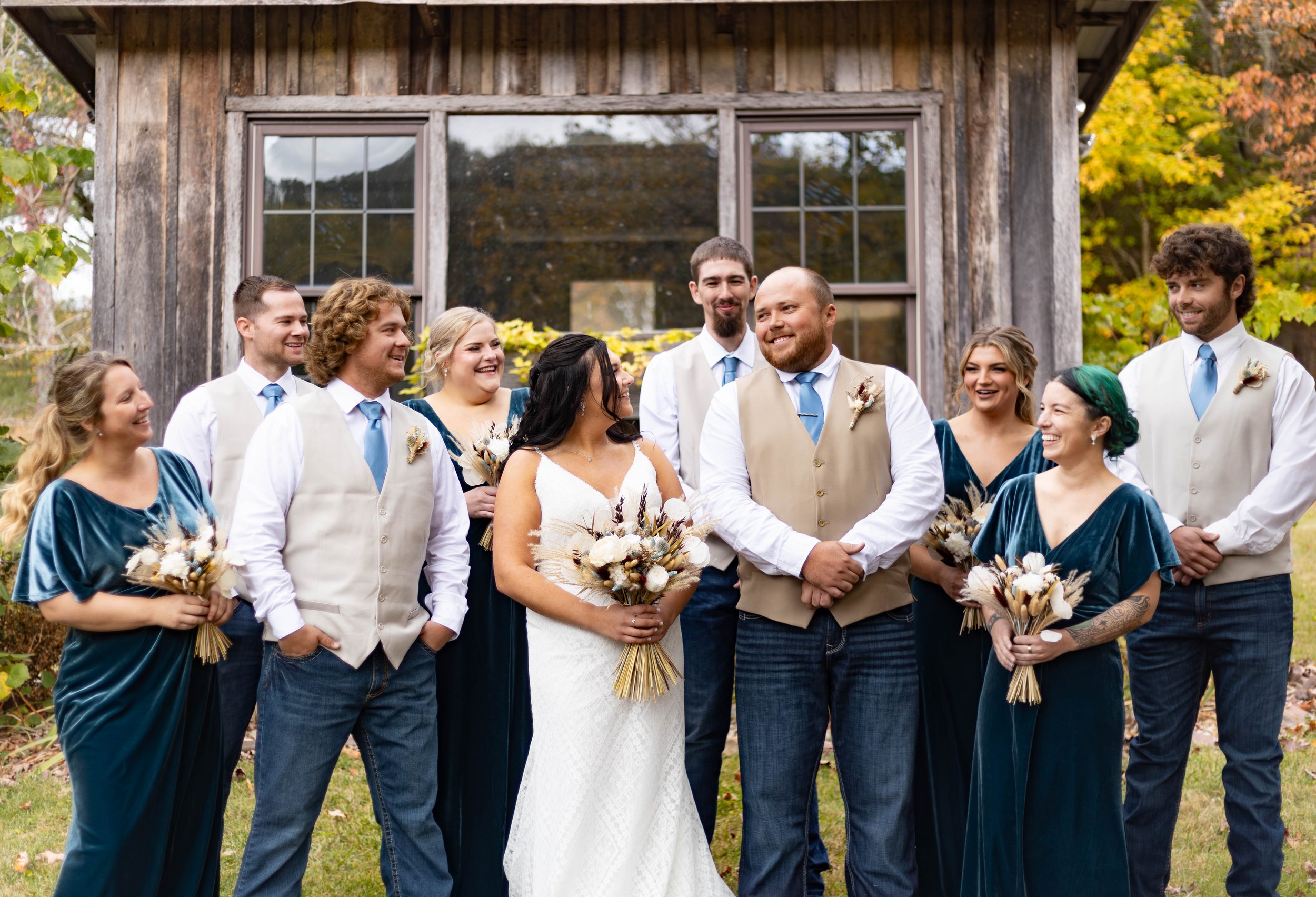 Group of eight people at a wedding, standing outdoors in front of a rustic wooden building, dressed in wedding attire with bouquets, smiling and talking.