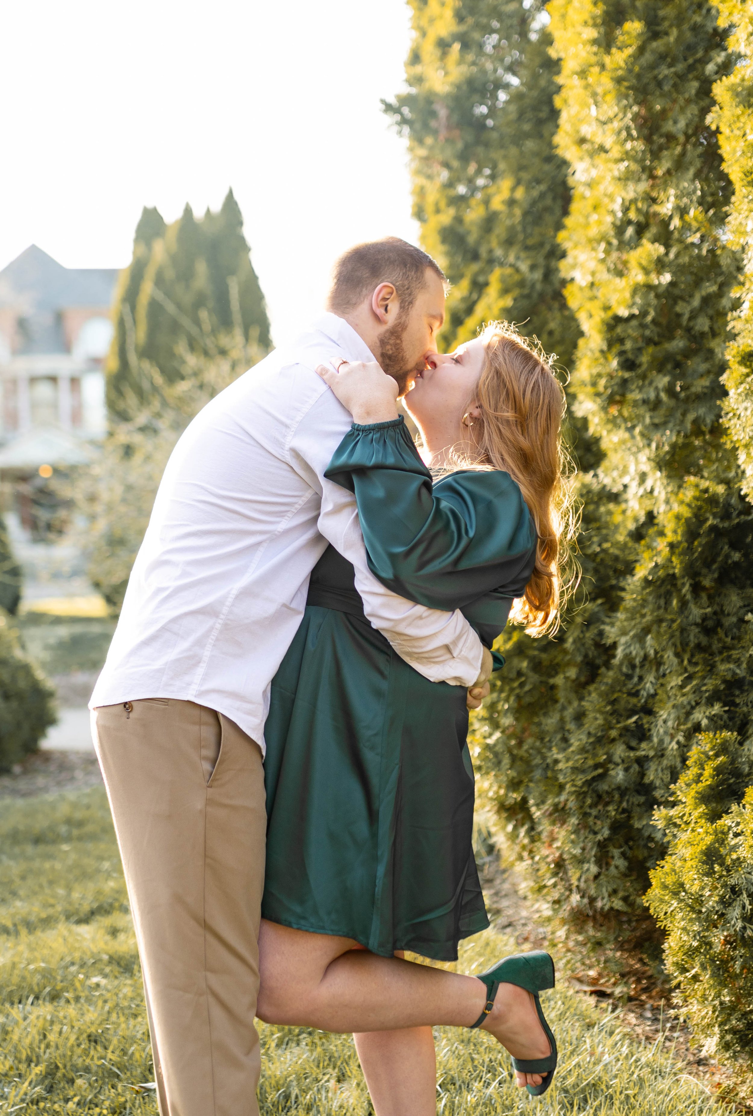 A couple kissing outdoors with greenery and trees in the background during golden hour.