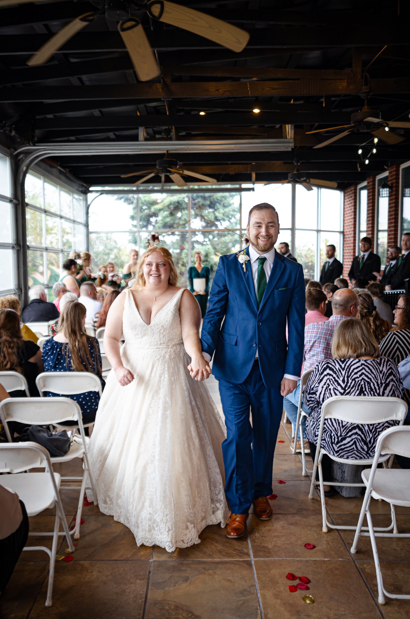 The bride and groom walking down the aisle holding hands during a wedding ceremony, with guests seated on either side and officiants in the background.