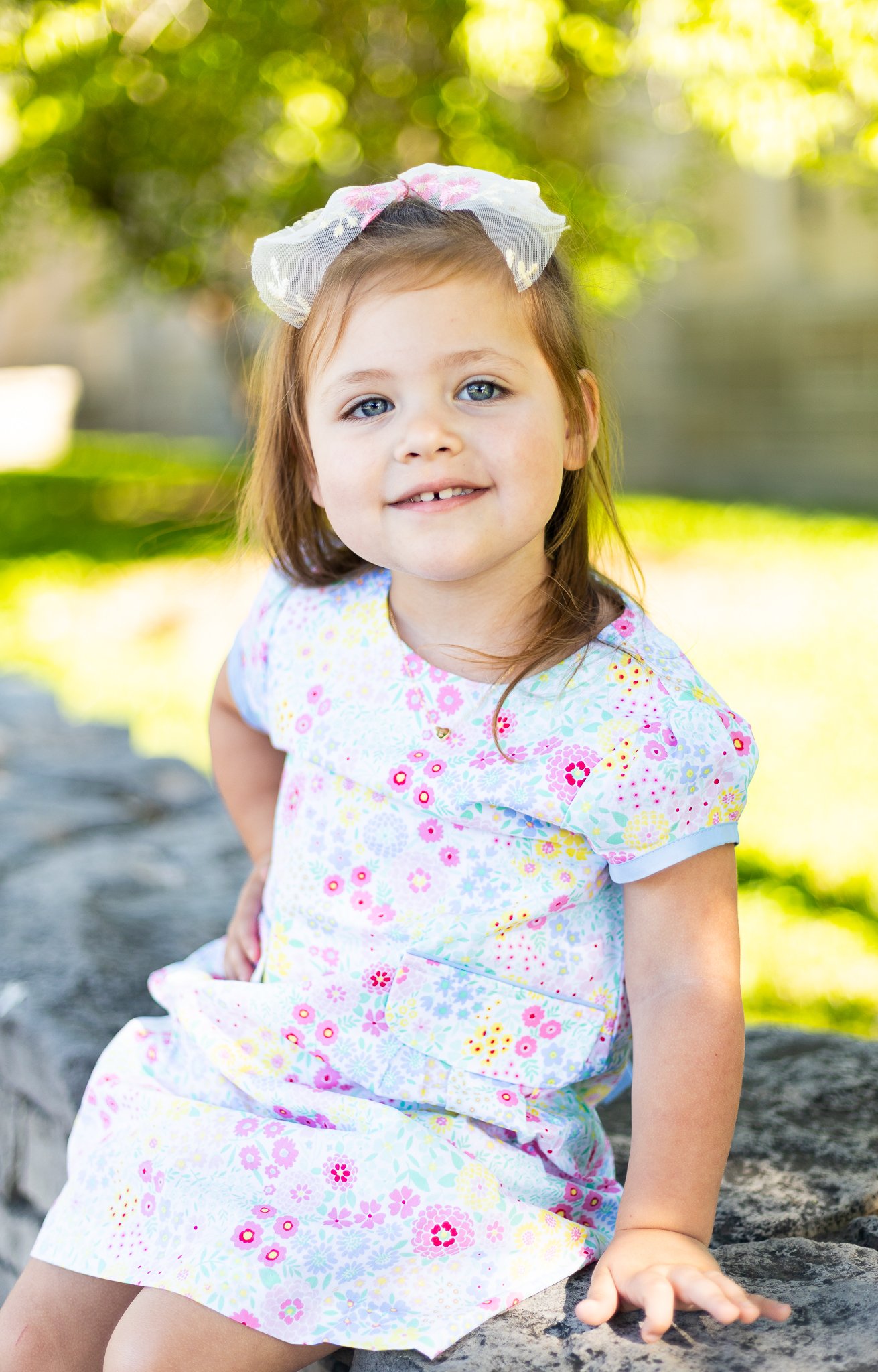 Young girl with light brown hair, blue eyes, and a bow in her hair, sitting on a stone ledge outdoors, wearing a colorful floral dress, with green trees and sunlight in the background.
