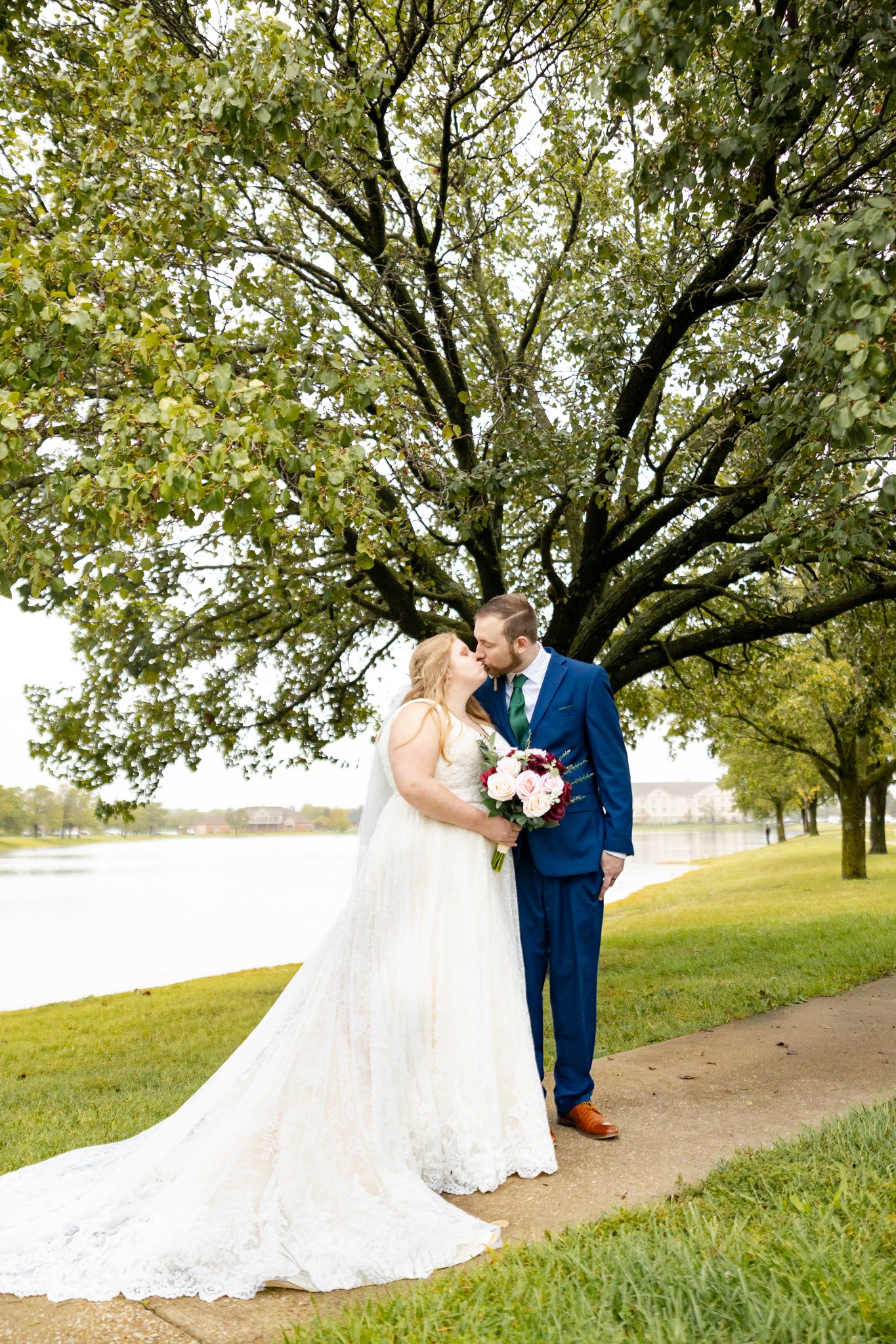A bride and groom sharing a kiss outdoors under a large tree near a lake, with the bride holding a bouquet of pink and white roses, on a cloudy day.