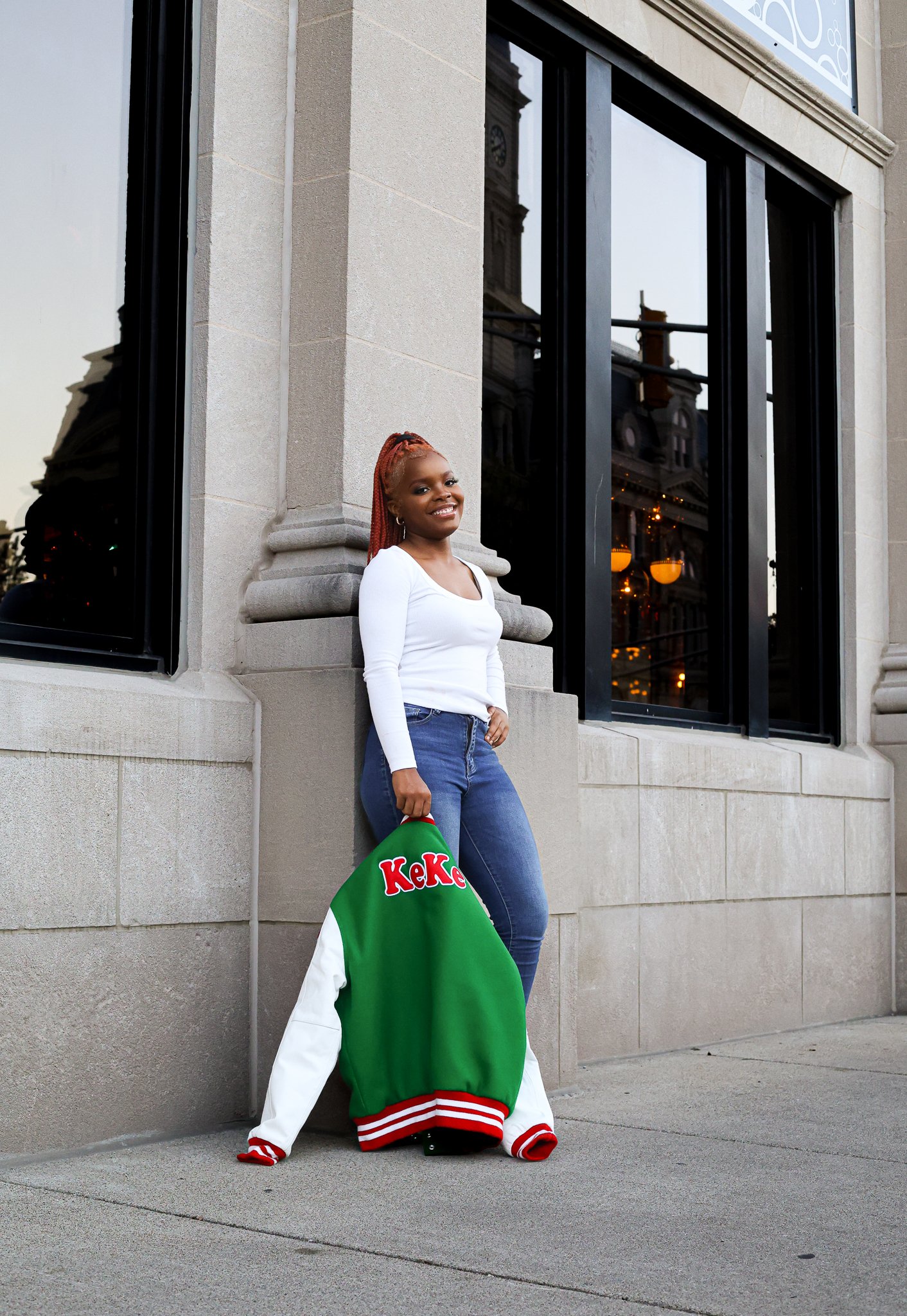 A young woman with braided hair, wearing a white long-sleeve shirt and blue jeans, stands against a stone building wall, smiling, holding a green and white college jacket with 'Keke' written on it.