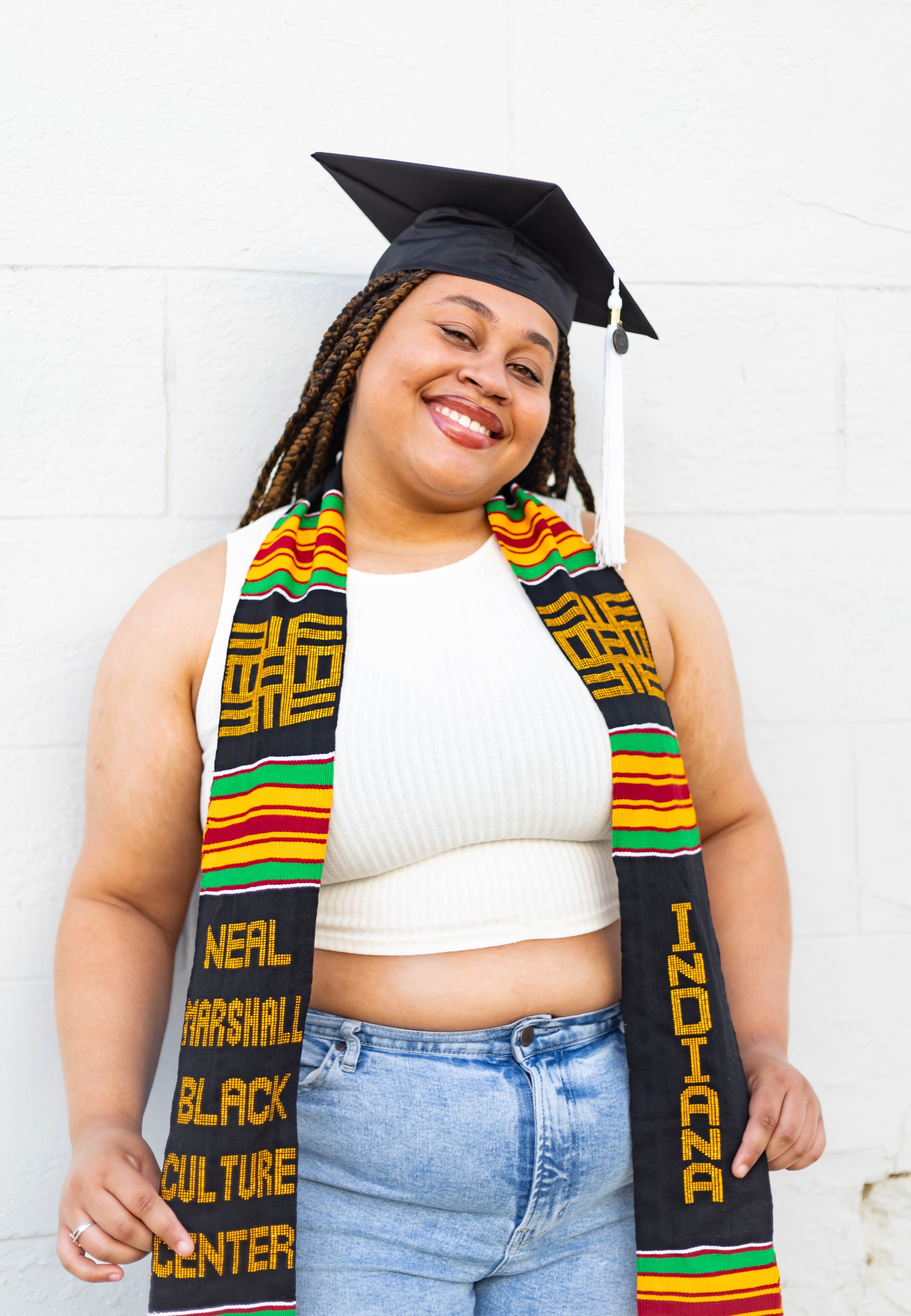 A young woman in graduation attire smiling against a white brick wall, wearing a black cap, a colorful Kente cloth stole, a white sleeveless top, and blue jeans.