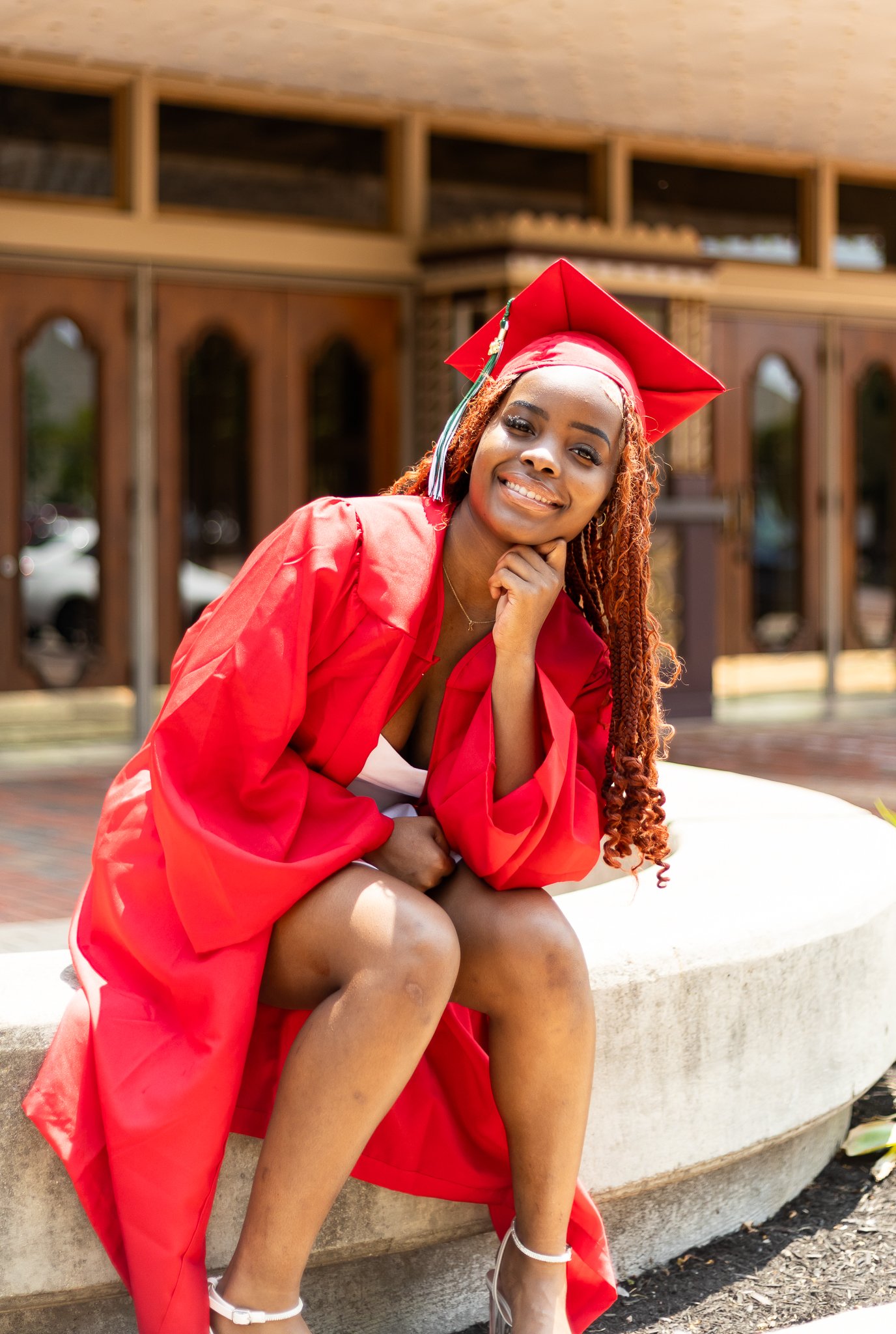 A young woman in a red graduation gown and cap sitting on a white stone bench, smiling and resting her chin on her hand outside a building with wooden doors.