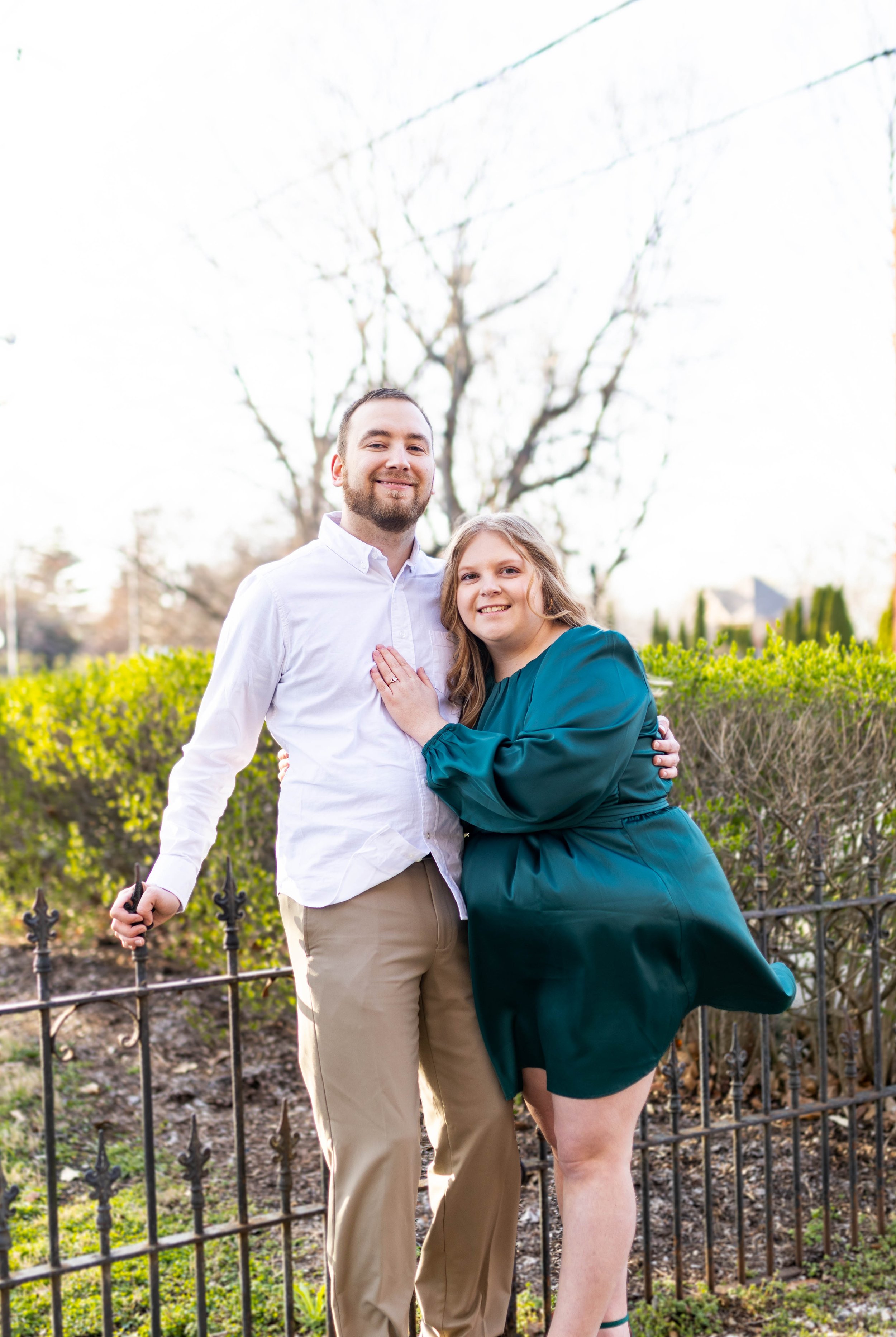 A couple smiling outdoors, with the woman leaning on the man who is holding a black object. They are standing near a small black metal fence, with green bushes and a leafless tree behind them.