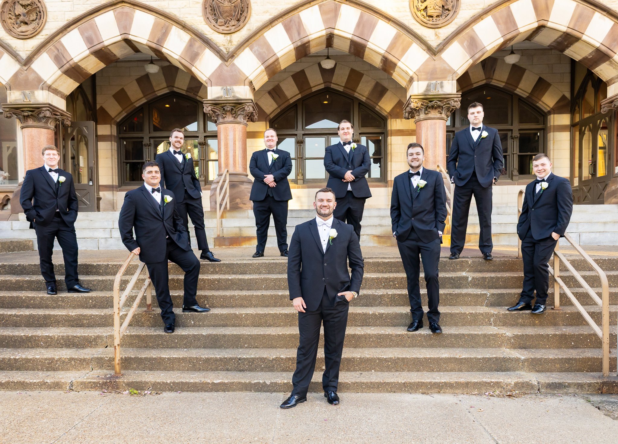 A group of ten men dressed in black tuxedos with white boutonnières, standing on steps in front of an ornate building, posing for a wedding photo.