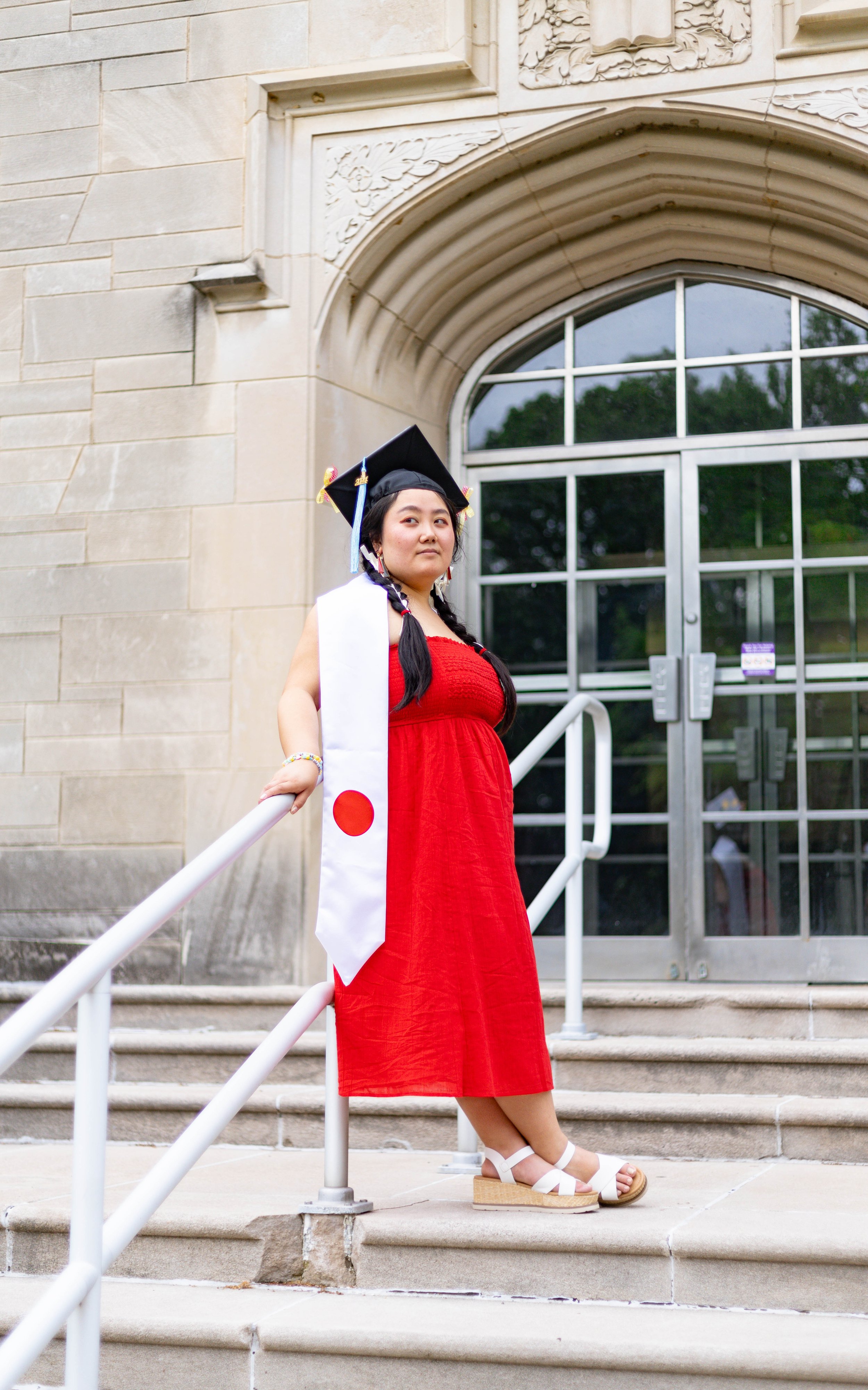 Young woman in a red dress and white sash with a Japanese flag, wearing a graduation cap, standing on steps outside a stone building with large glass doors.