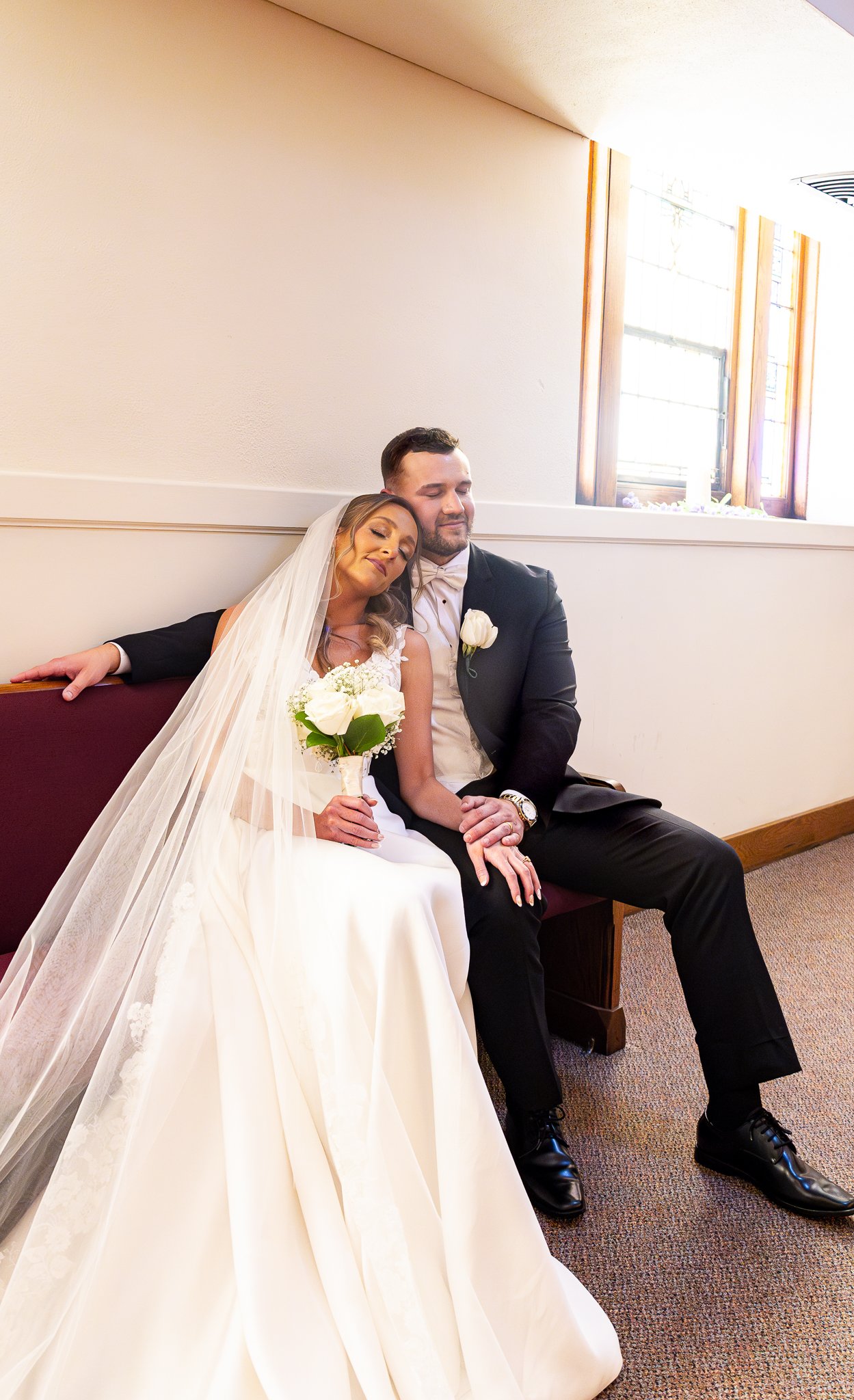 Bride and groom sitting close together on a bench, with the bride resting her head on the groom's shoulder, both with peaceful expressions, bride holding a bouquet of white roses, groom wearing a black tuxedo with a white boutonniere, in a room with