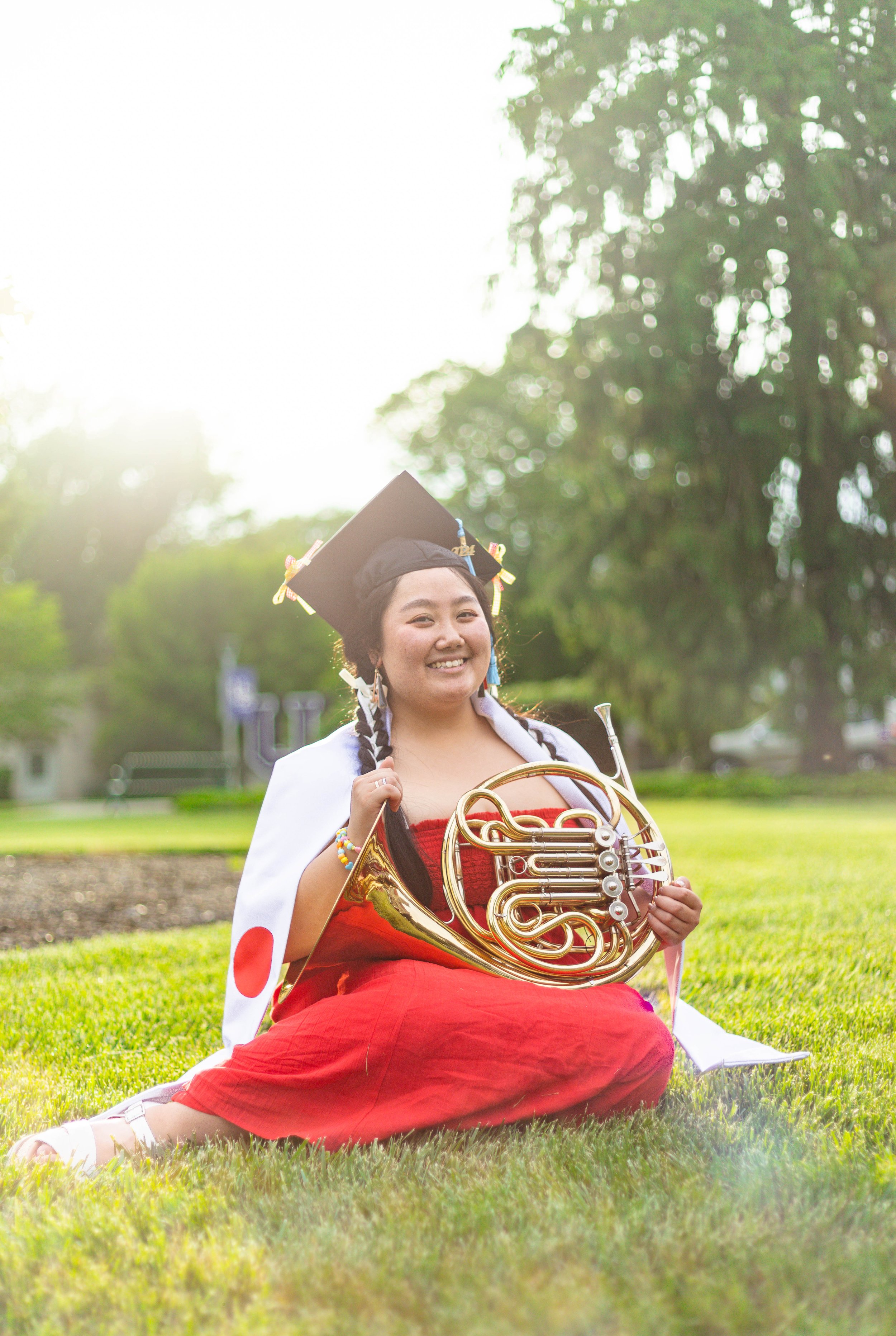 Young woman in a red dress, graduation cap, and white cape with a Japanese flag, sitting on grass in a park, holding a french horn, smiling with sunlight shining behind her.