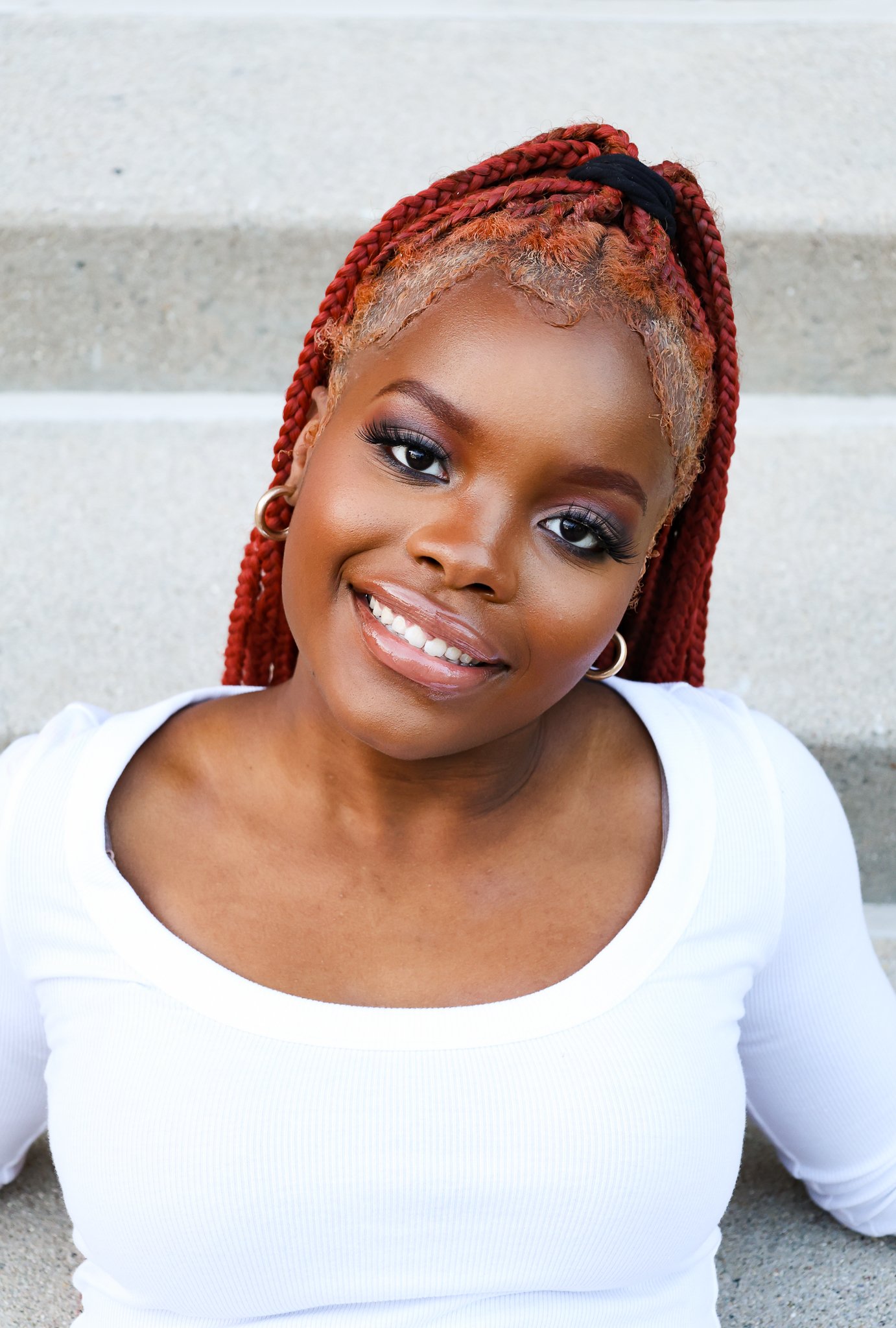 Close-up of a smiling woman with red braids, hoop earrings, and wearing a white top, sitting against a concrete wall.