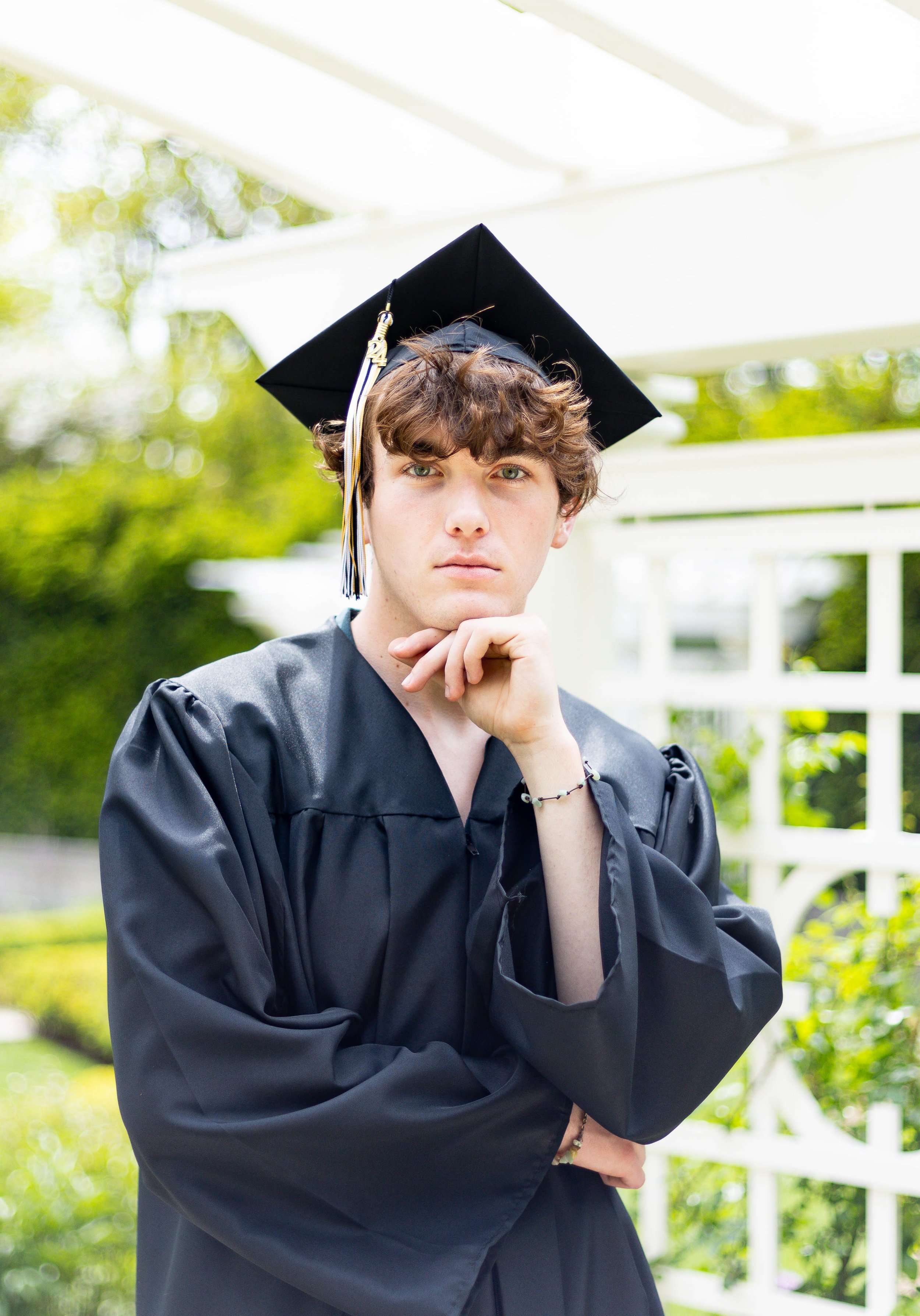 A young man in a black graduation gown and cap outdoors, posing with his chin resting on his hand, in front of a white lattice and green foliage.