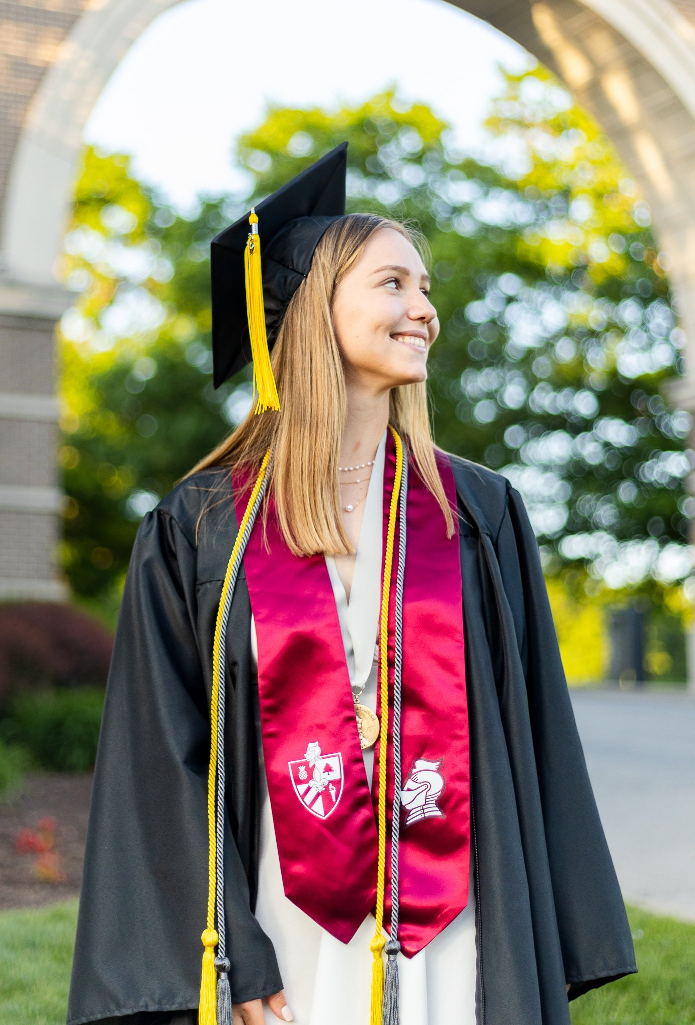 A young woman in black graduation cap and gown with a maroon and gold stole standing outdoors, smiling and looking to the right.