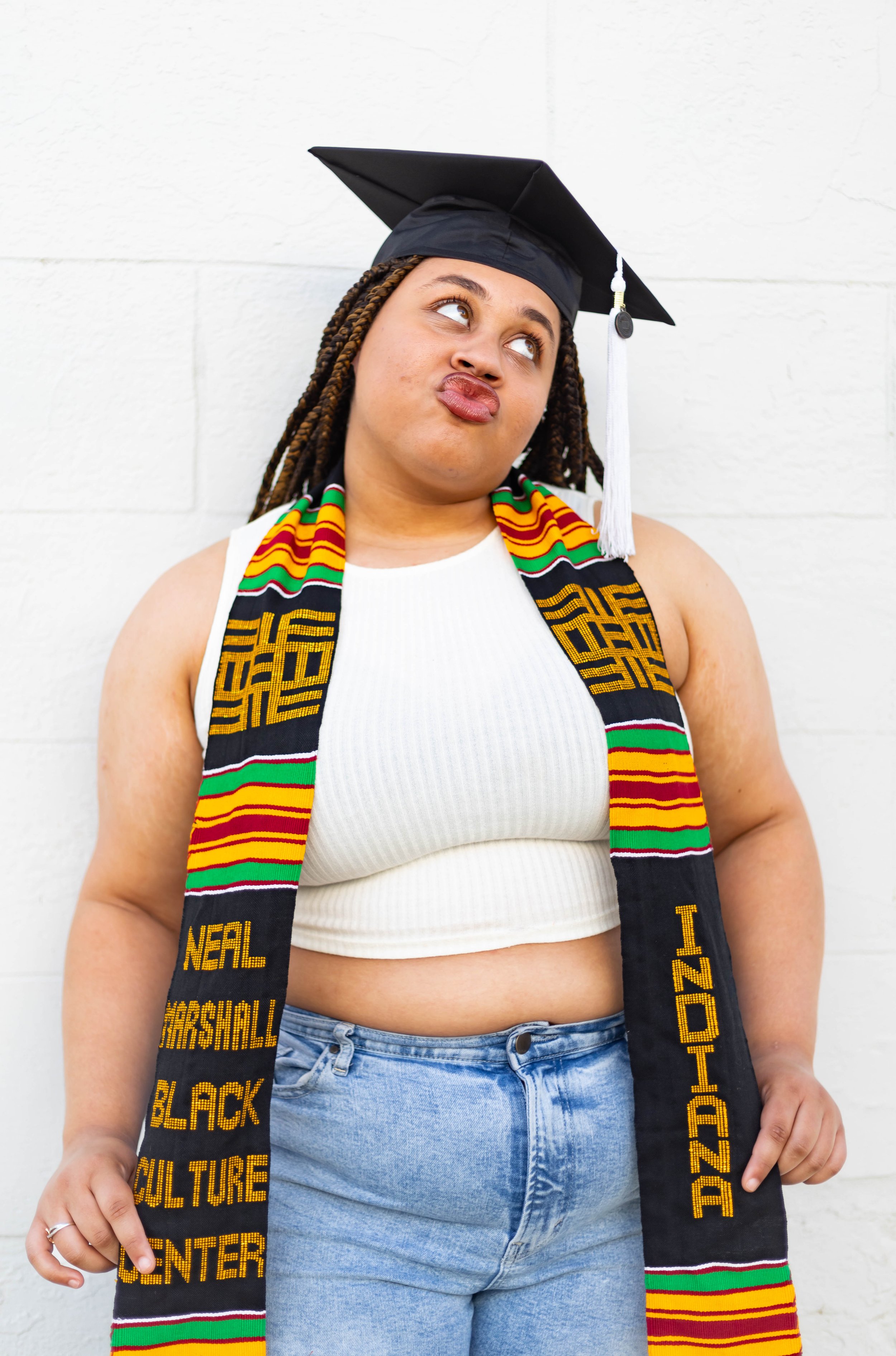 Young woman with braided hair wearing a graduation cap, white crop top, blue jeans, and a colorful kente cloth stole, standing against a white brick wall, making a playful face.