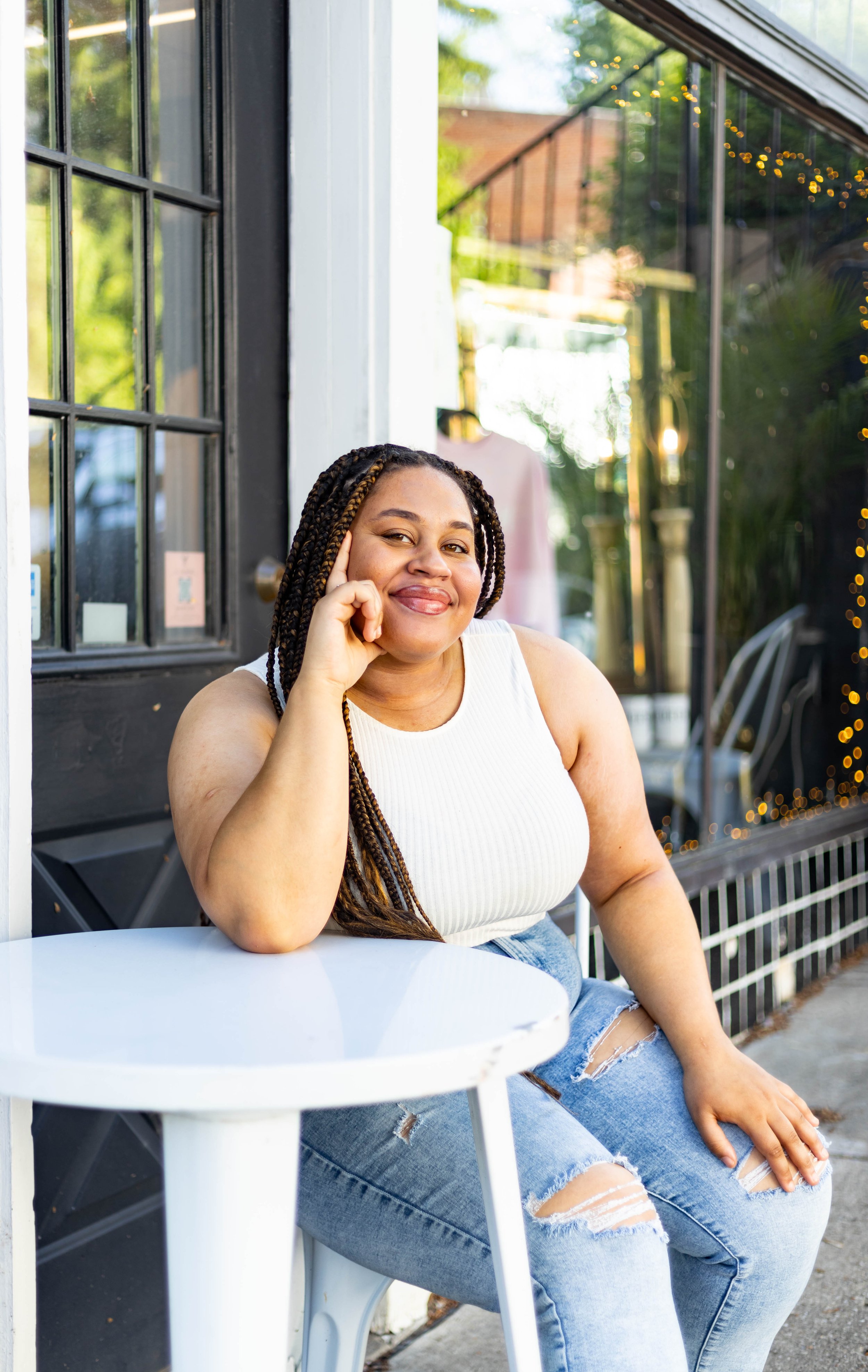A woman with braided hair, wearing a white tank top and ripped jeans, sitting outside at a white table, smiling, with her elbow resting on the table and her hand touching her temple.