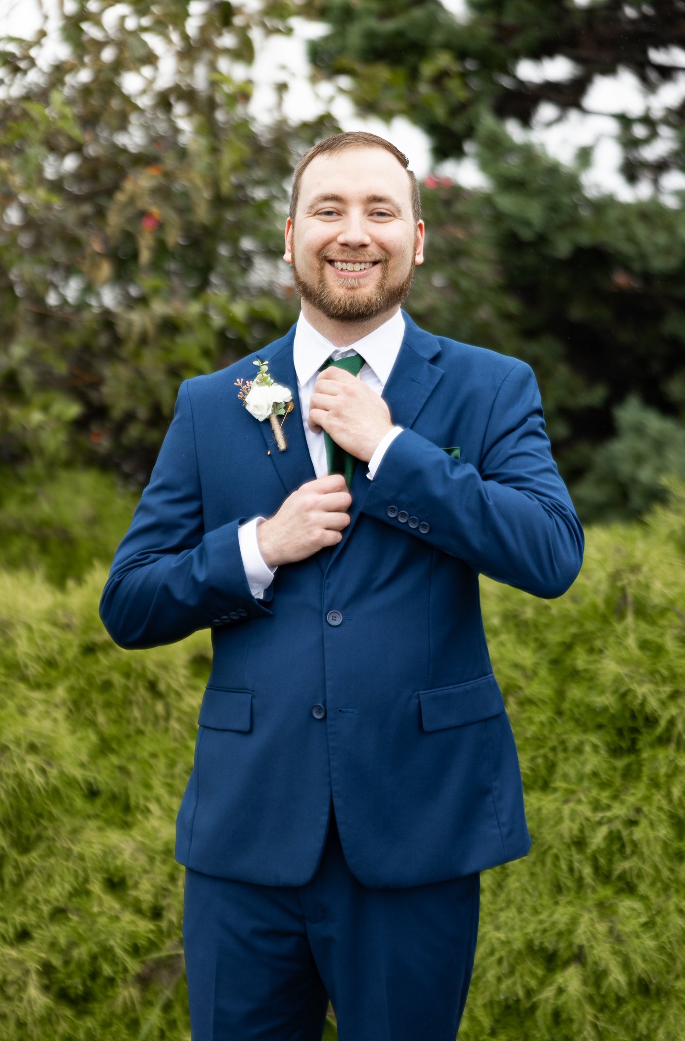 A man in a blue suit adjusting his tie outdoors, with greenery and trees in the background.