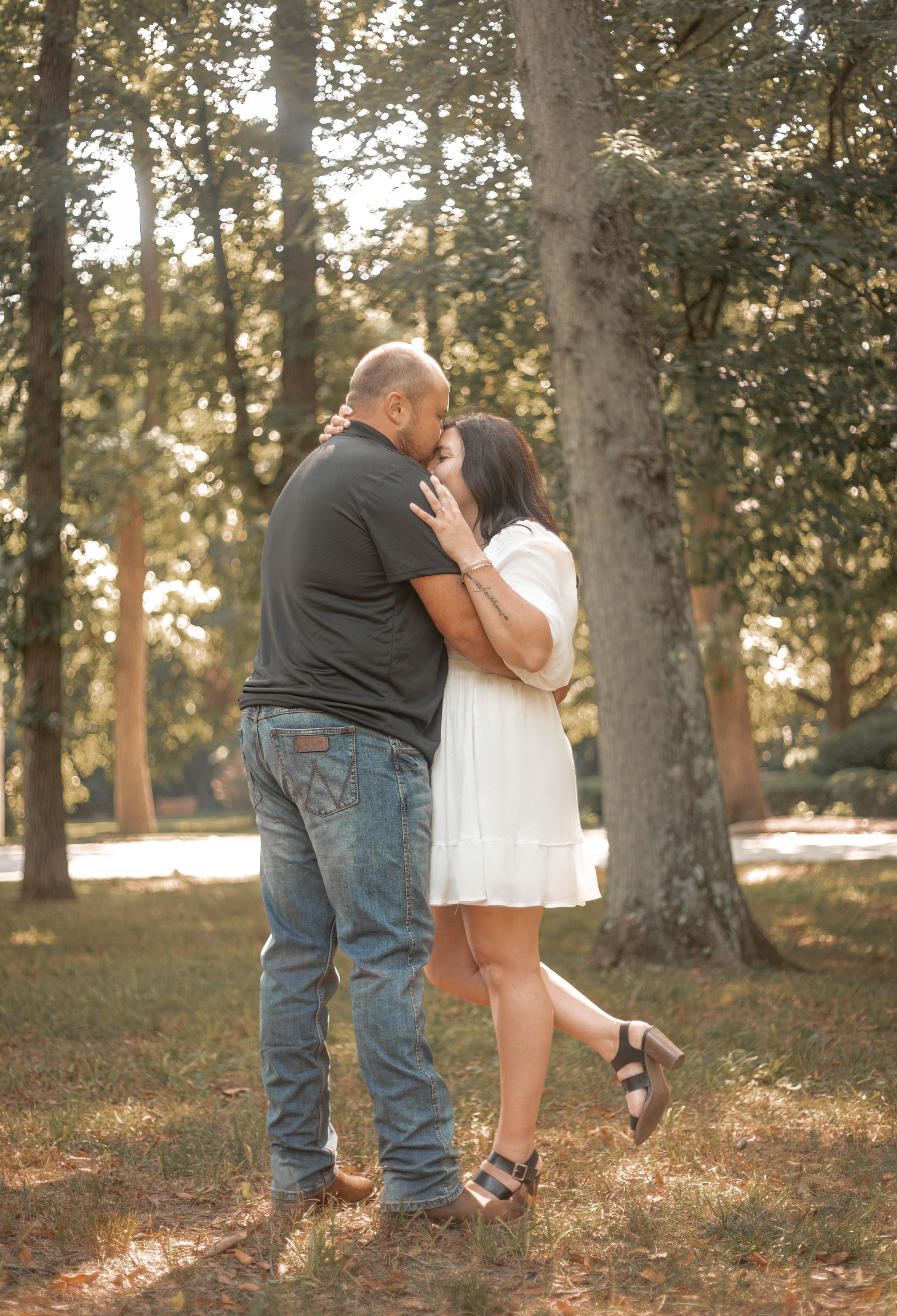 A couple sharing a kiss in a park during sunset, surrounded by large trees and sunlight filtering through the leaves.