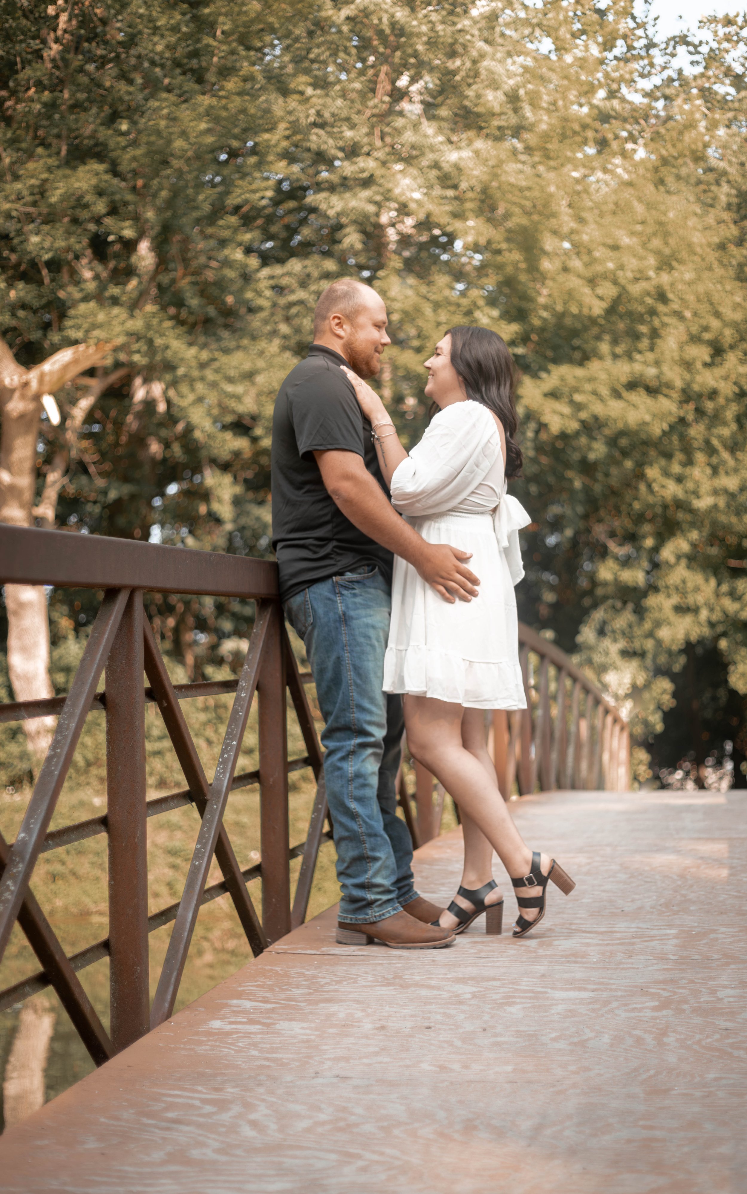 A couple stands close on a wooden bridge with green trees in the background, smiling at each other.
