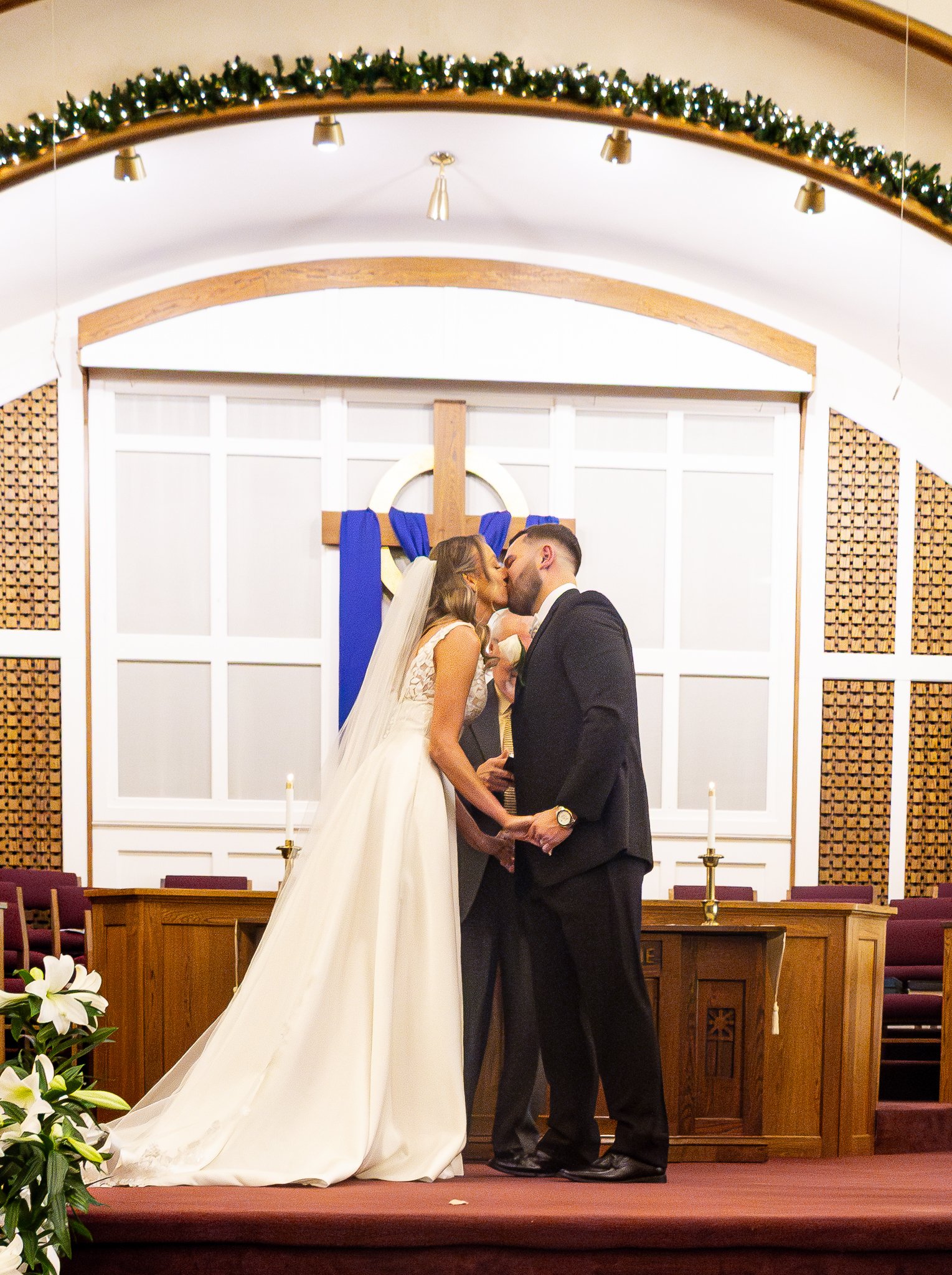 A bride and groom kissing during their wedding ceremony in a church, with an altar decorated with a wooden cross, candles, and floral arrangements.