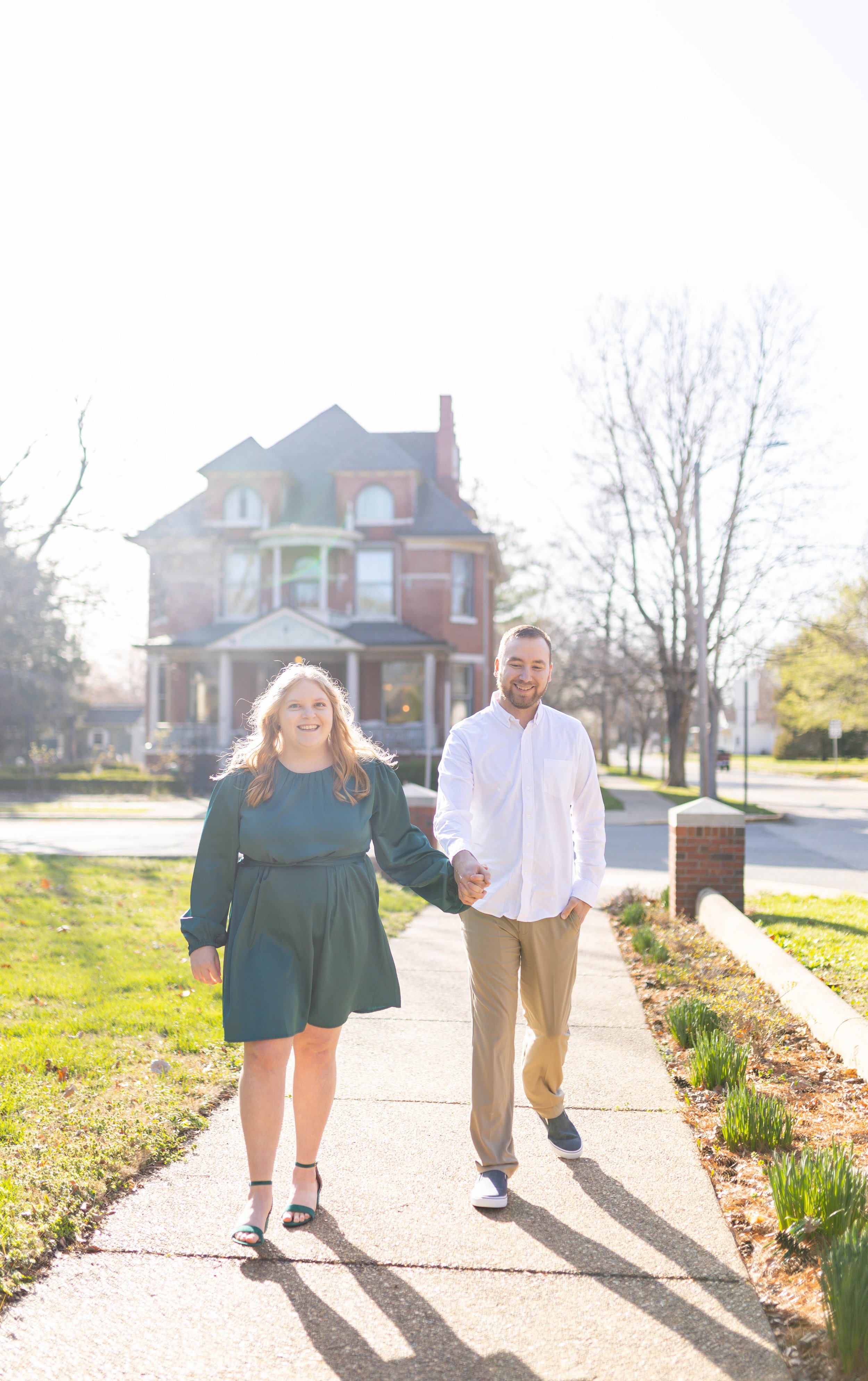 A couple walking hand-in-hand on a sidewalk in front of a large Victorian-style house, smiling and enjoying a sunny day.