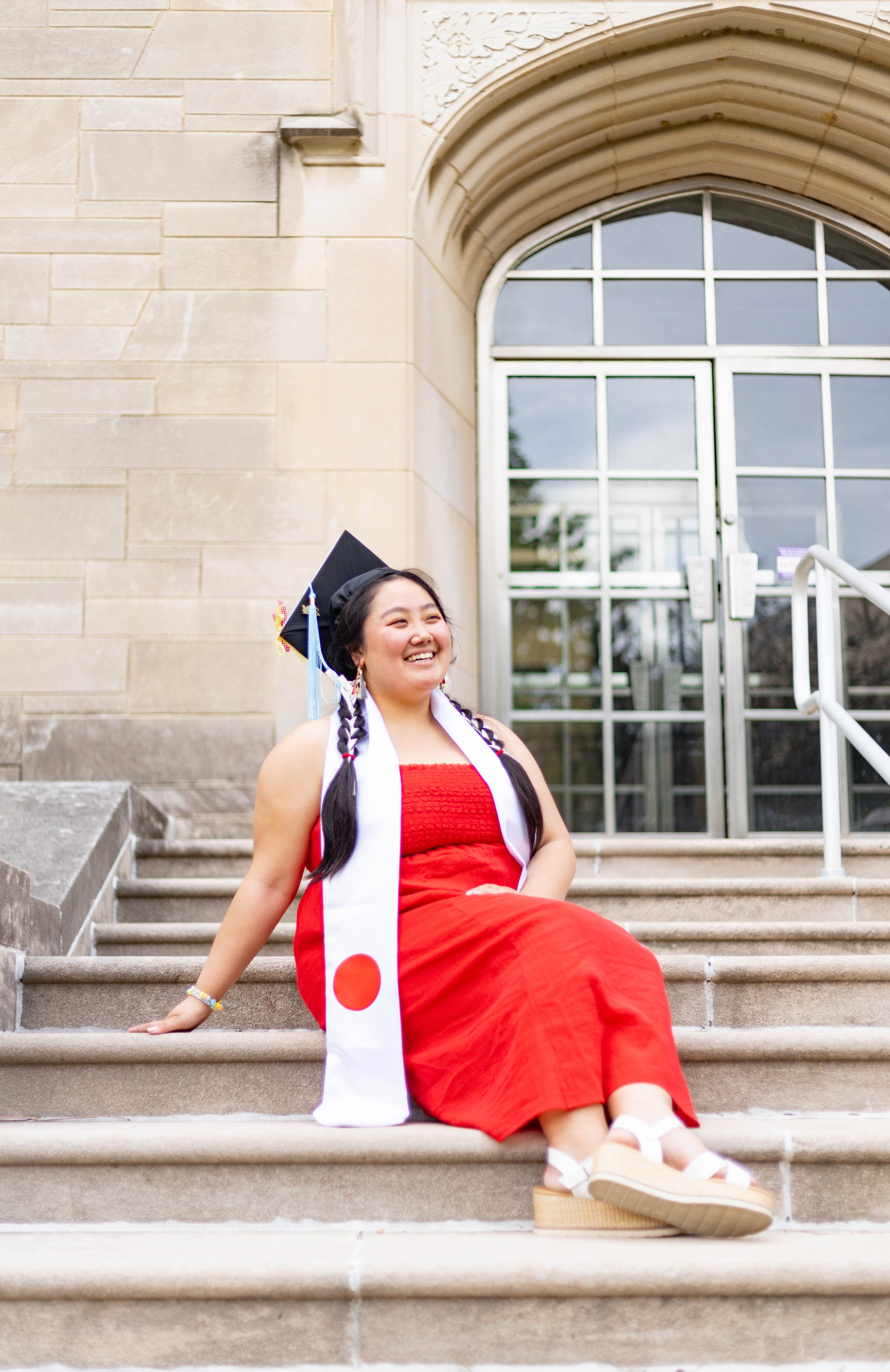 A young woman wearing a red dress, a graduation cap, and a white stole with a red circle, sitting on the steps in front of a large building with a glass door.