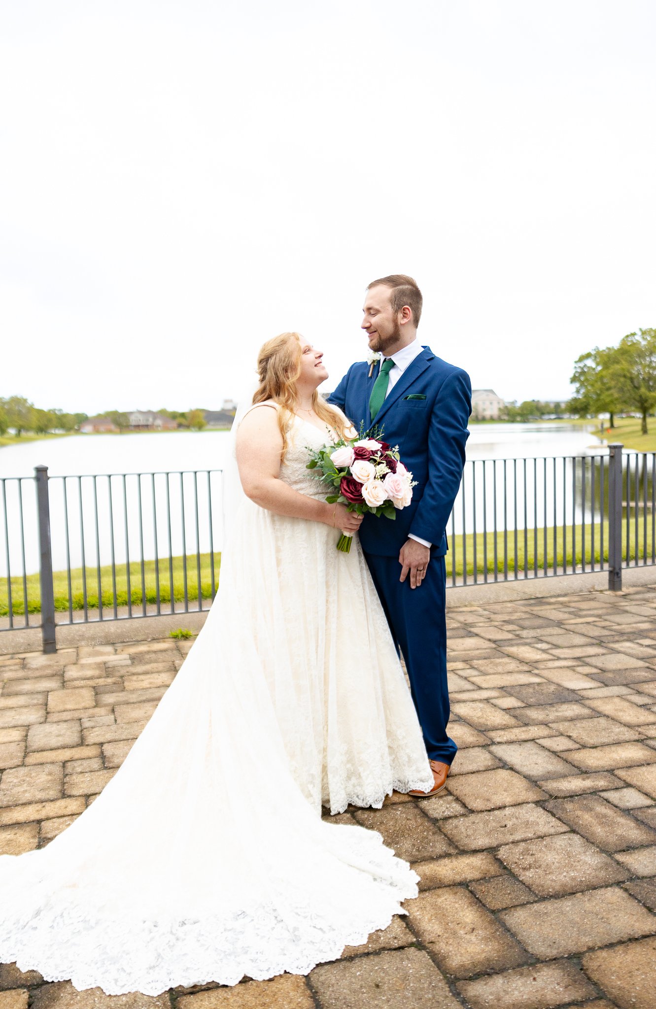 A bride and groom standing by a lake on their wedding day, with the bride holding a bouquet of flowers and looking at the groom, who is smiling back at her.