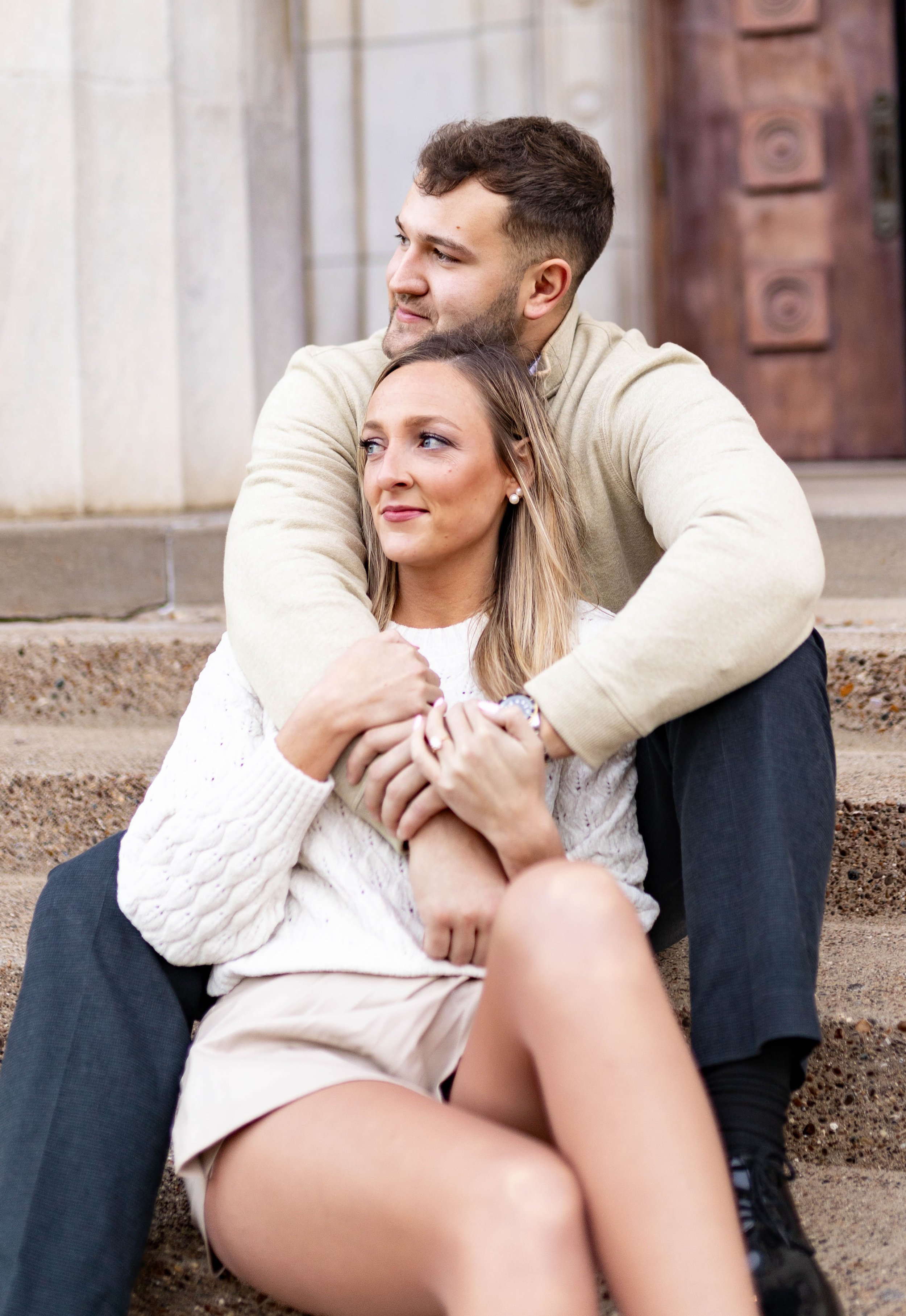 A young couple sitting on concrete steps outside a building, with the man embracing the woman from behind. The woman looks attentive and content, while the man gazes off into the distance with a gentle smile.