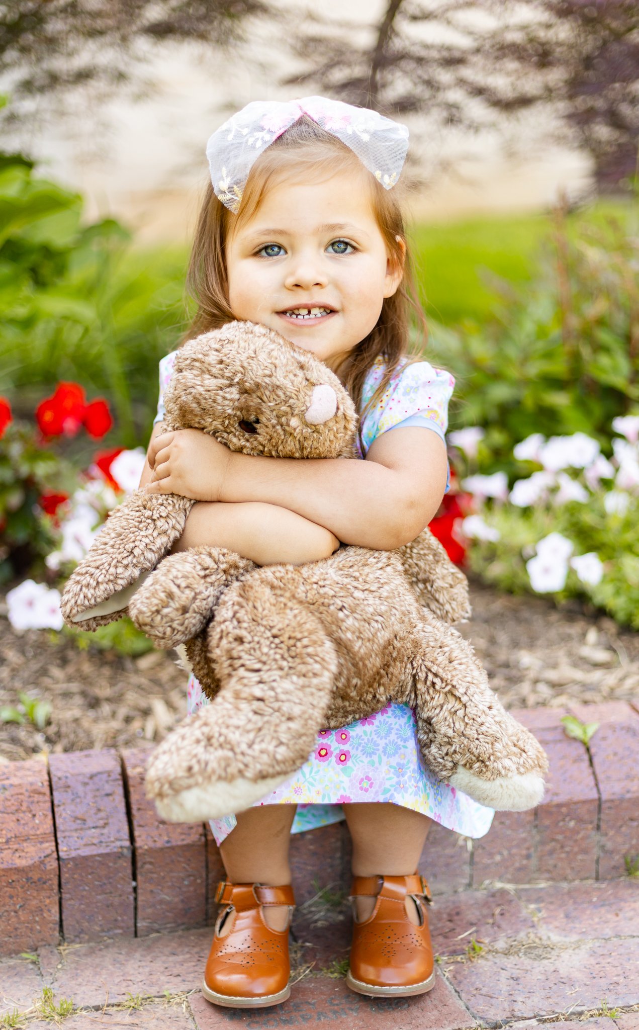 A young girl with blue eyes and light brown hair, wearing a white dress with colorful flowers, tan shoes, and a pink bow headband, is hugging a large plush teddy bear outdoors near a flower bed.