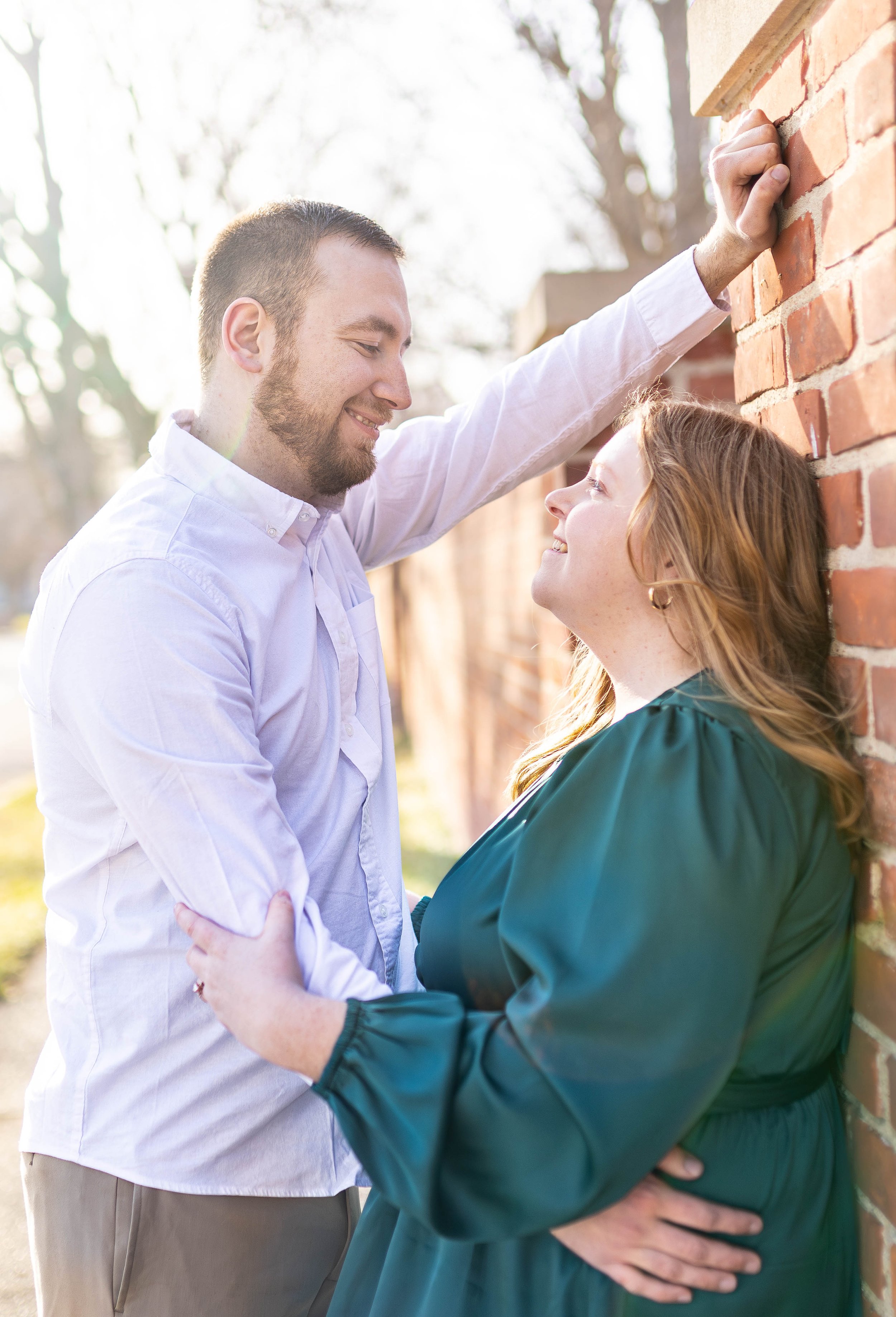 A man leaning against a brick wall outdoors, looking lovingly at a woman who is smiling up at him, with sunlight filtering through trees in the background.