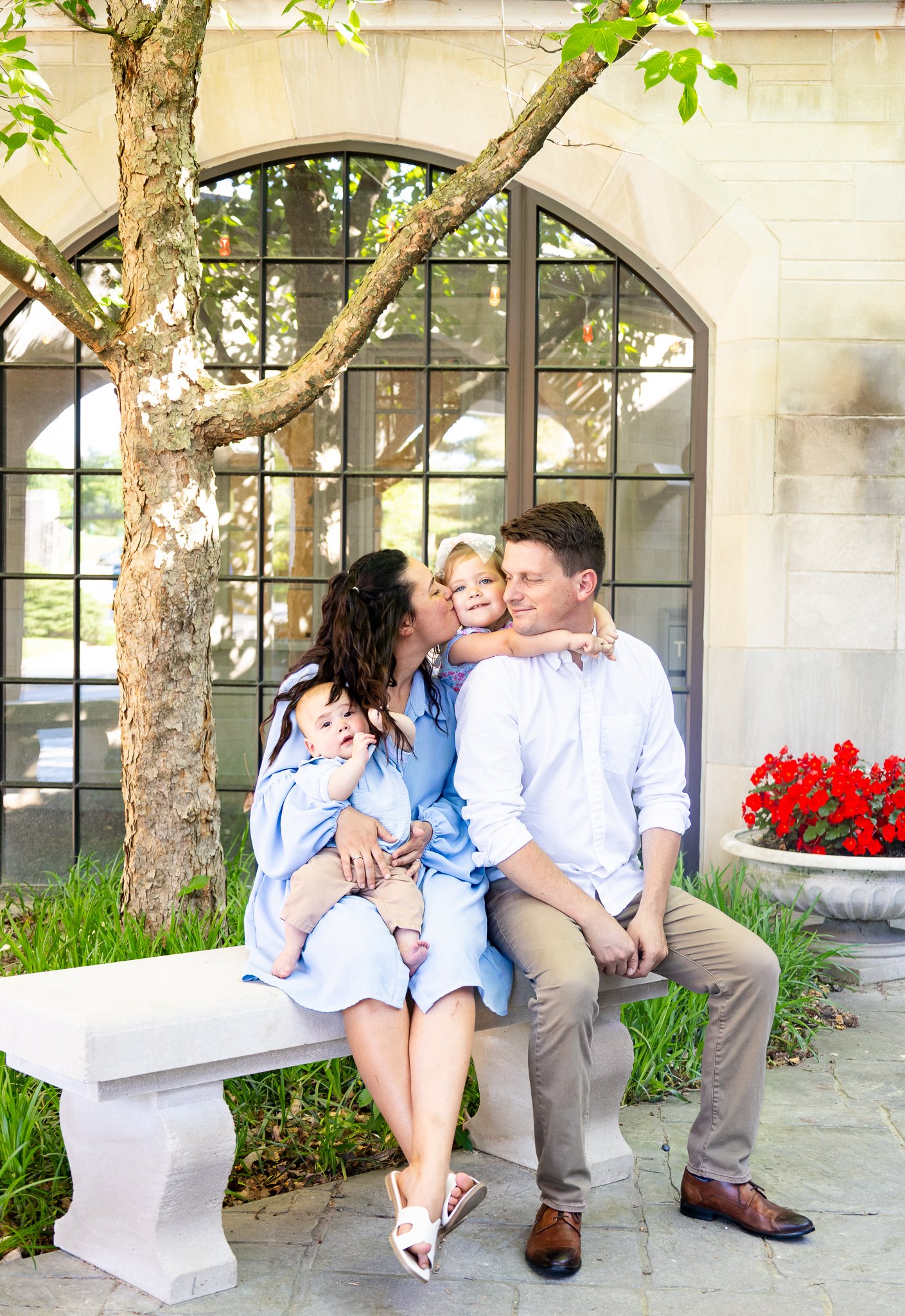 A family of four sitting on a bench outdoors in front of a building with large window panes. The woman is kissing one of the children, a young girl who is hugging her, while the man is sitting with a baby on his lap. There is a tree with green leaves