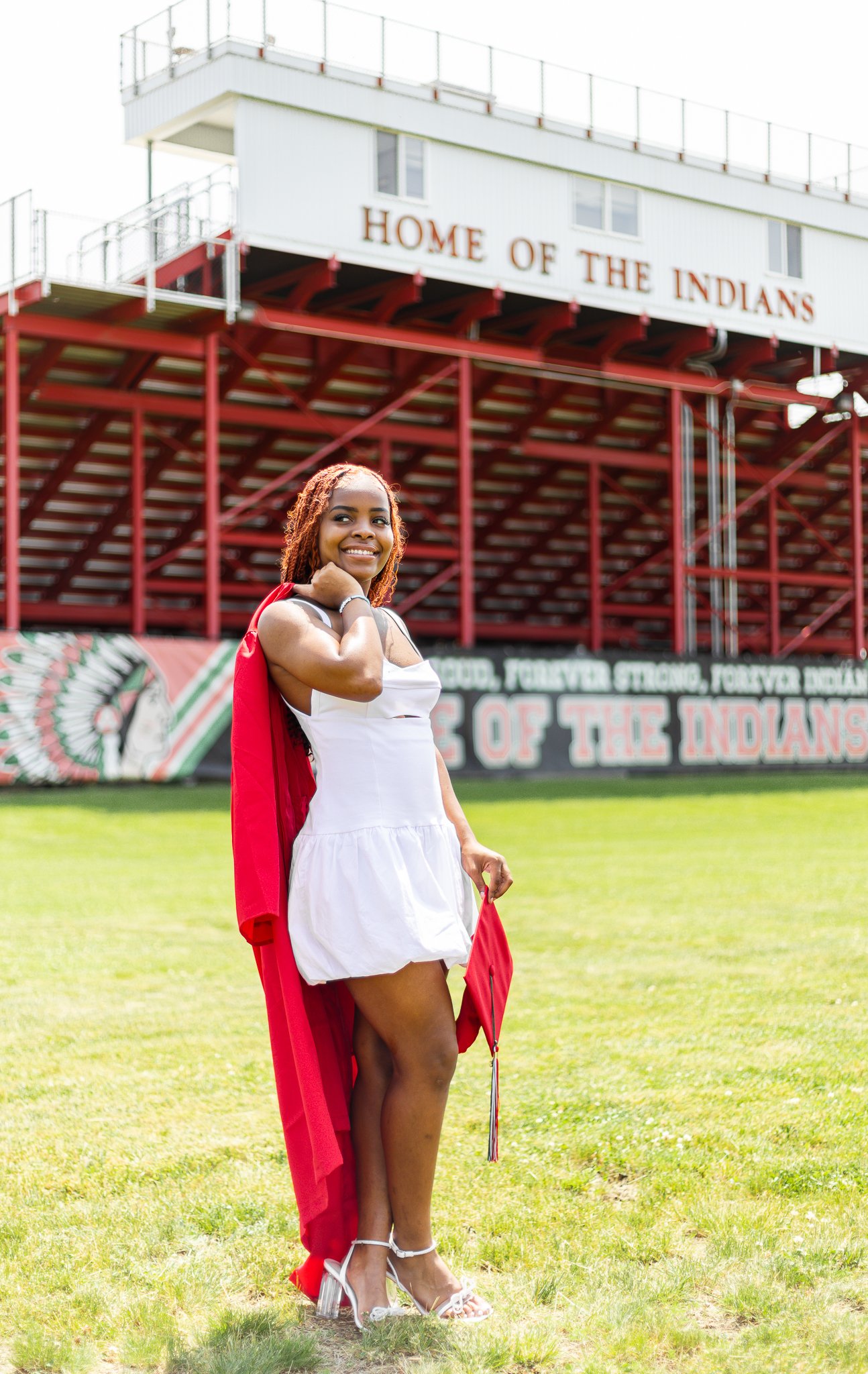 A woman in a white dress with a red graduation gown draped over her shoulders, holding a diploma, standing on a grassy field in front of a football stadium with the sign "Home of the Indians" and a banner that reads "Forever Strong, Forever Indians".