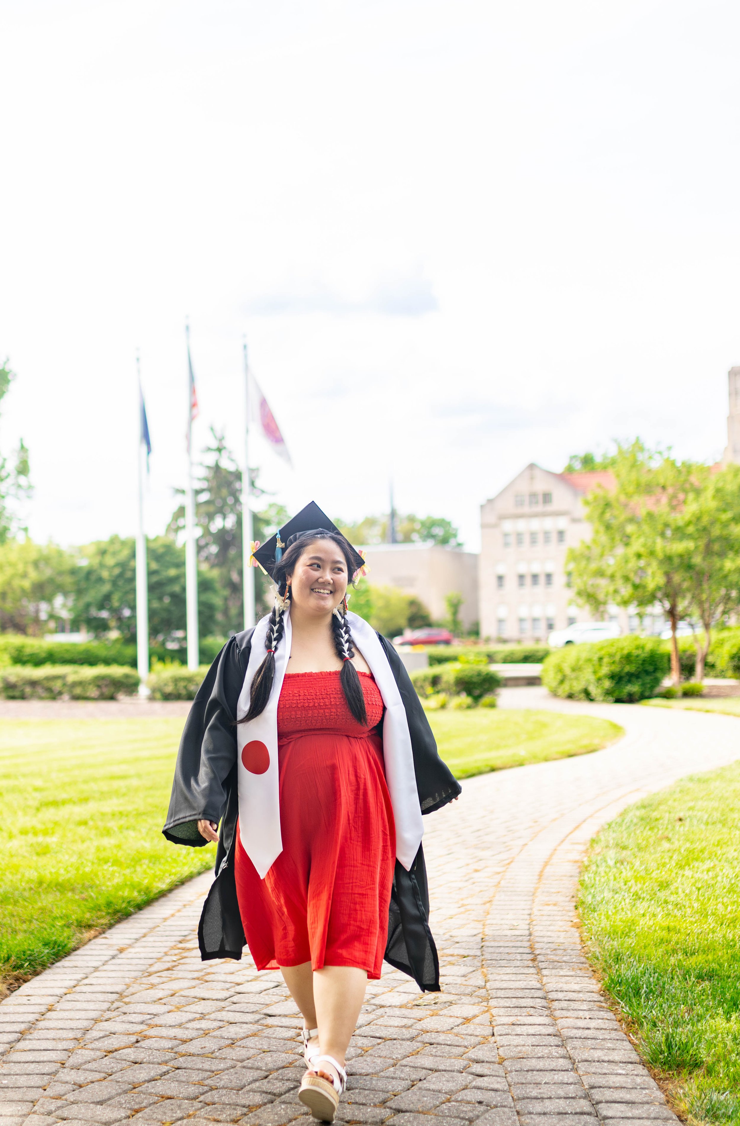 A young woman in a graduation cap and gown walking on a brick path outdoors, smiling, with a university campus and flags in the background.