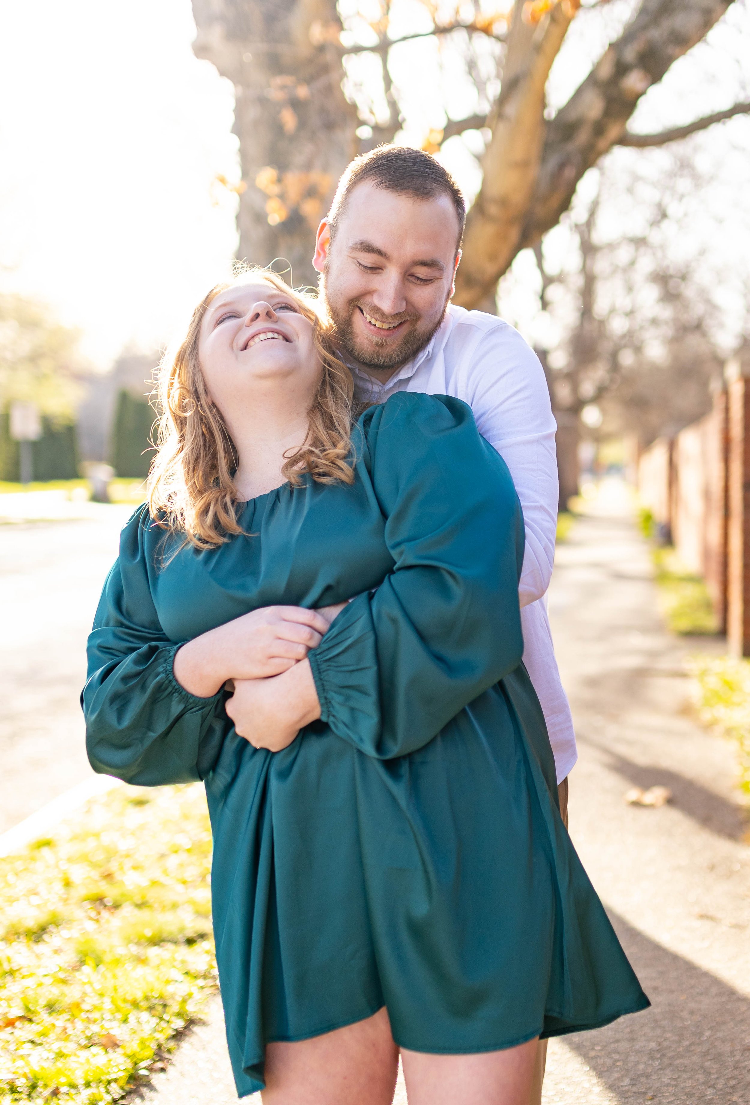 A man and woman enjoying a joyful moment outdoors on a sunny day, with trees and a sidewalk in the background. The woman is wearing a teal dress and is smiling with her head tilted back, while the man embraces her from behind, smiling down at her.