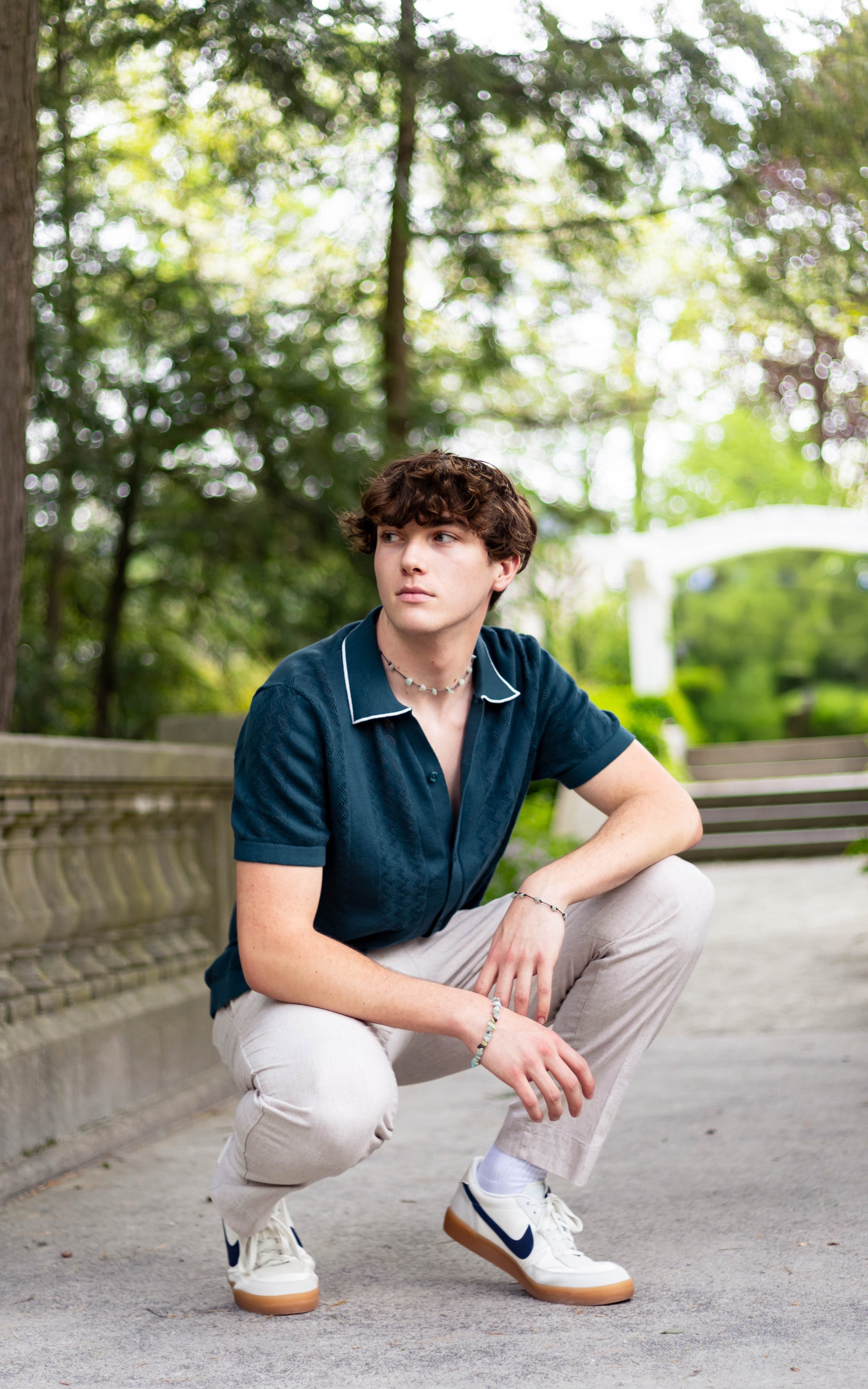 A young man with curly hair crouching outdoors on a paved walkway, wearing a dark teal short-sleeved shirt, beige pants, white sneakers with navy swoosh, and jewelry in a park with trees and benches in the background.