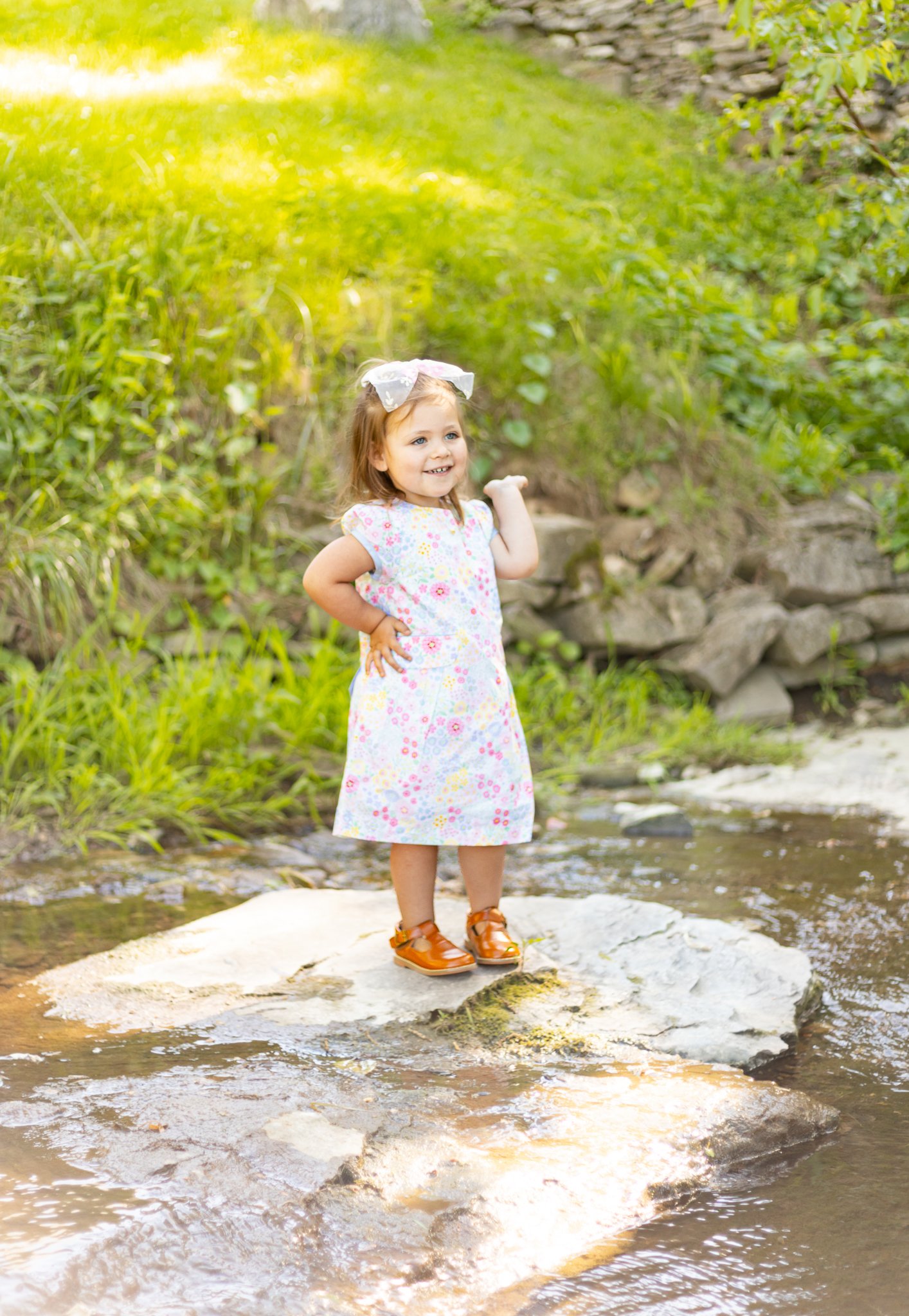 A young girl with a white bow in her hair, wearing a floral dress and brown shoes, stands on a large rock in a creek, smiling and posing with one hand on her hip in a lush, green outdoor setting.
