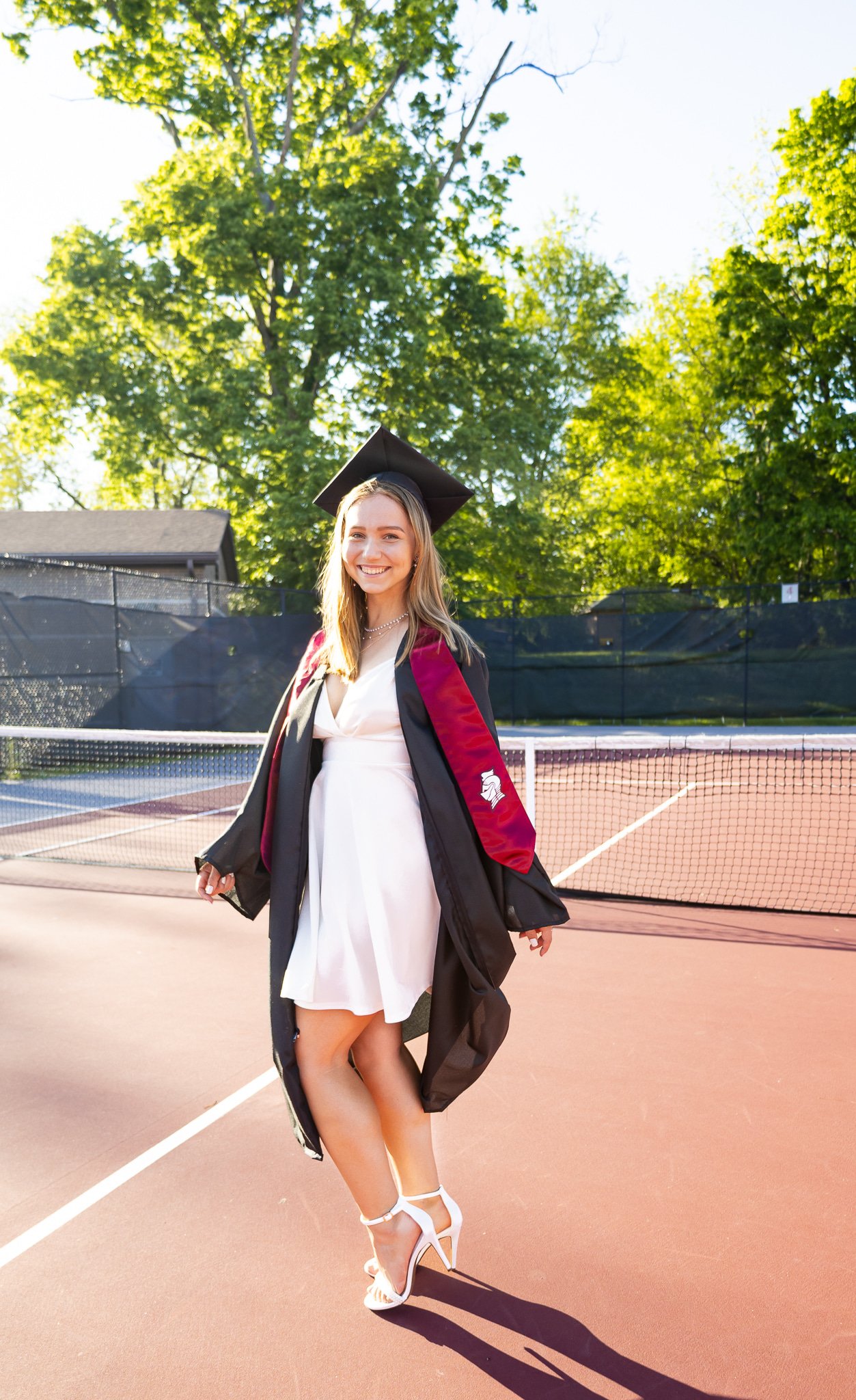 A young woman in a white dress and high heels is celebrating her graduation outdoors on a tennis court, wearing a black graduation cap and gown with maroon accents and a white emblem on the sleeve.