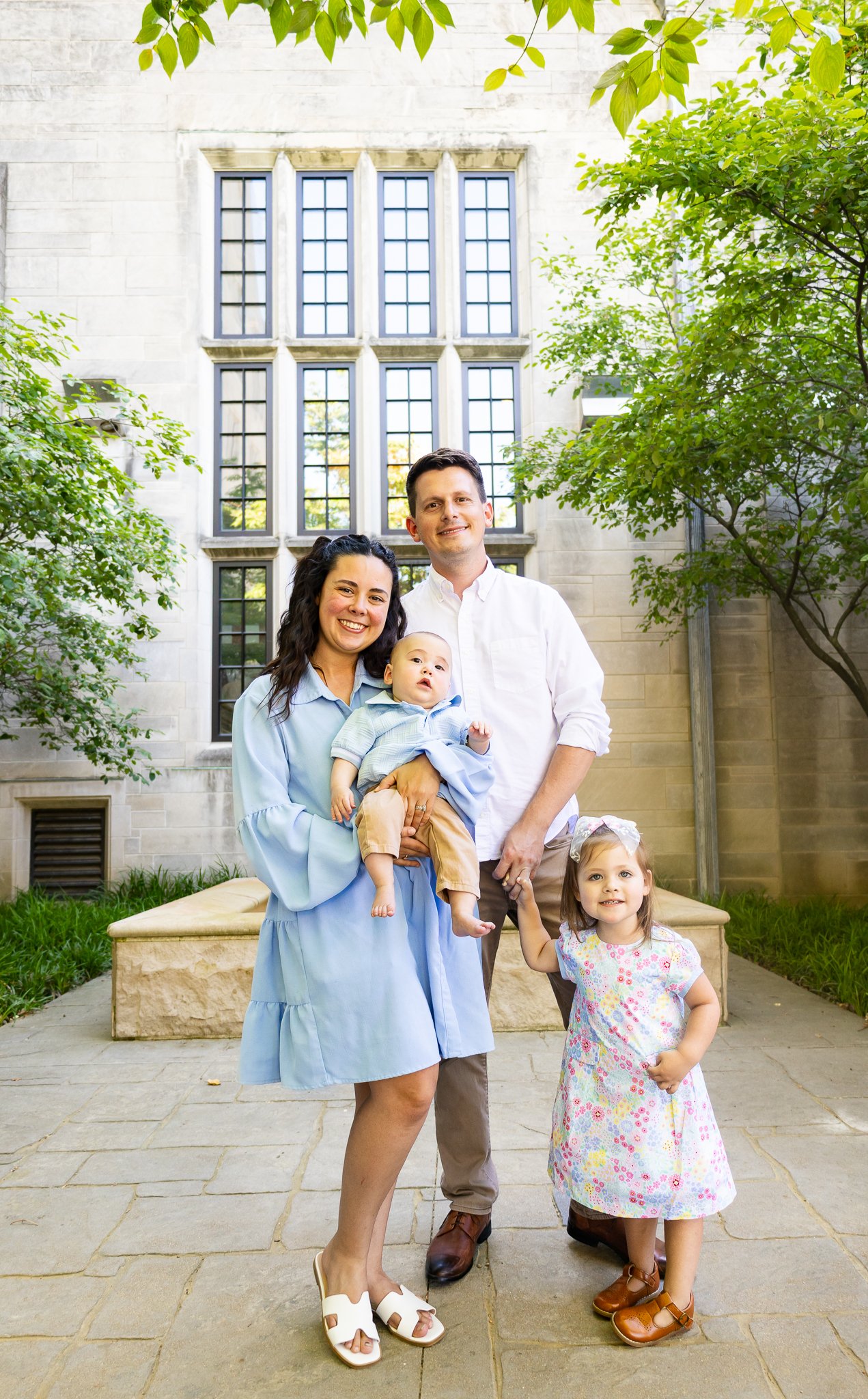 A family of four standing outdoors in front of a stone building with large windows, surrounded by green trees and grass. The mother holds a baby boy, the father is smiling, and a young girl with a bow in her hair is holding the father's hand.