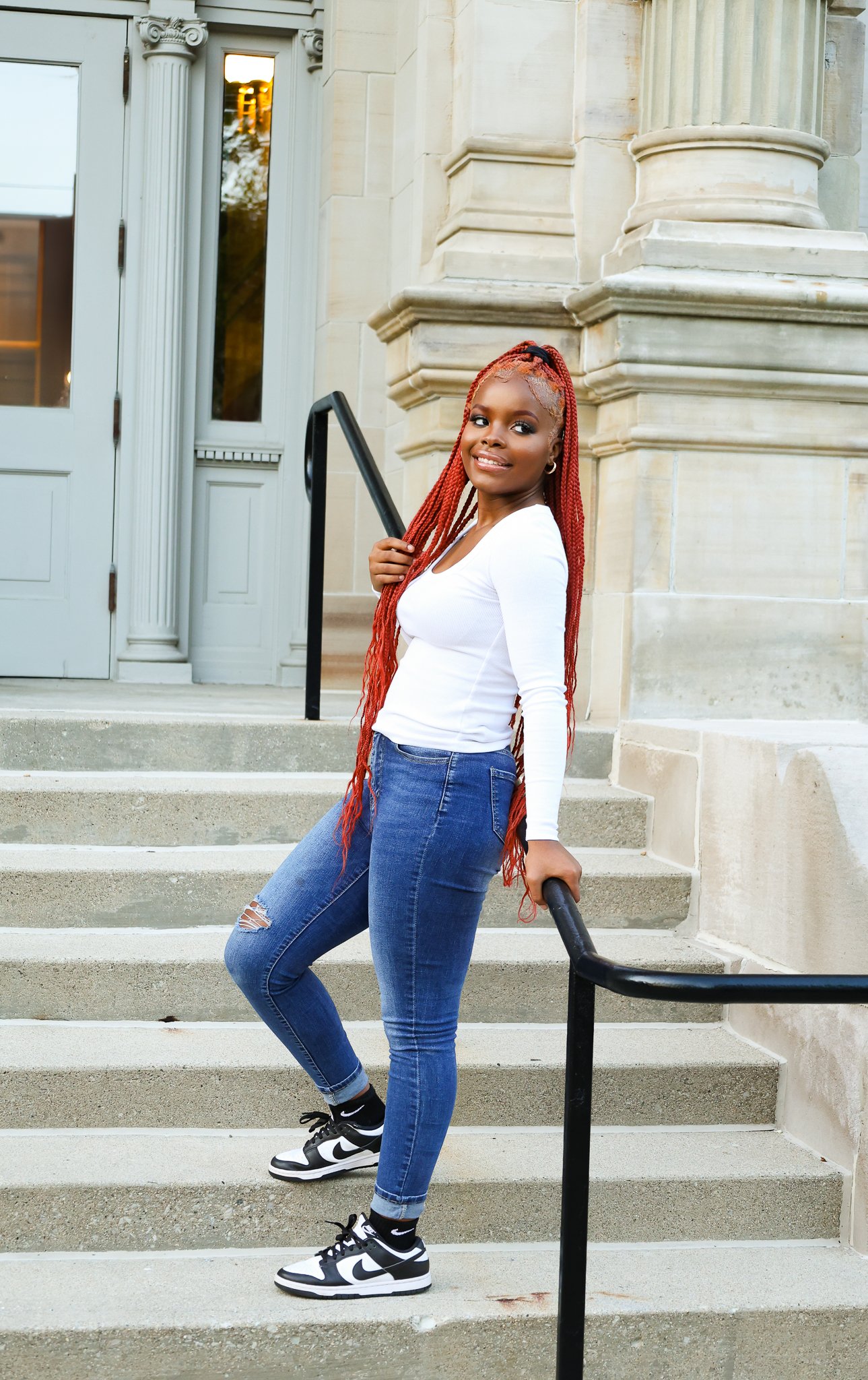 A young woman with long red braids, wearing a white long-sleeve shirt, ripped jeans, and Nike sneakers, standing on outdoor stairs in front of a historic building with columns and an arched entrance, smiling at the camera.