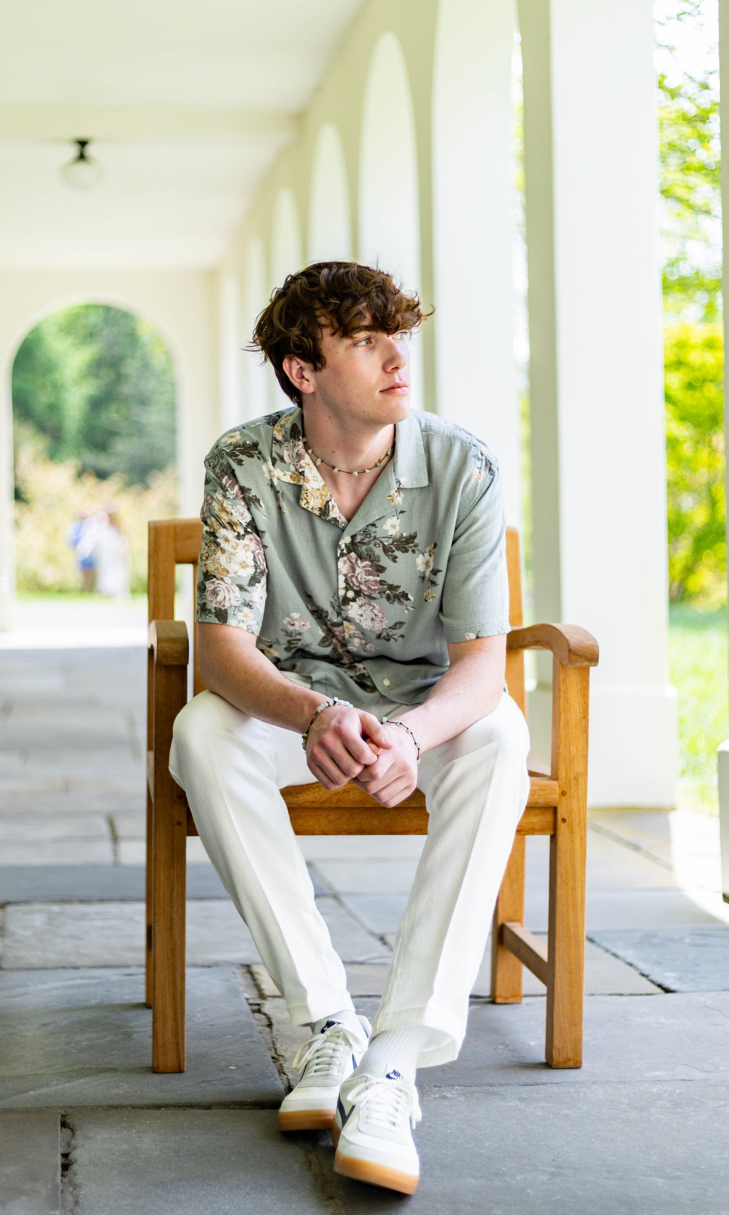 A young man with curly hair sitting on a wooden chair on a covered porch with a white archway background and green outdoor scenery, looking to his left.