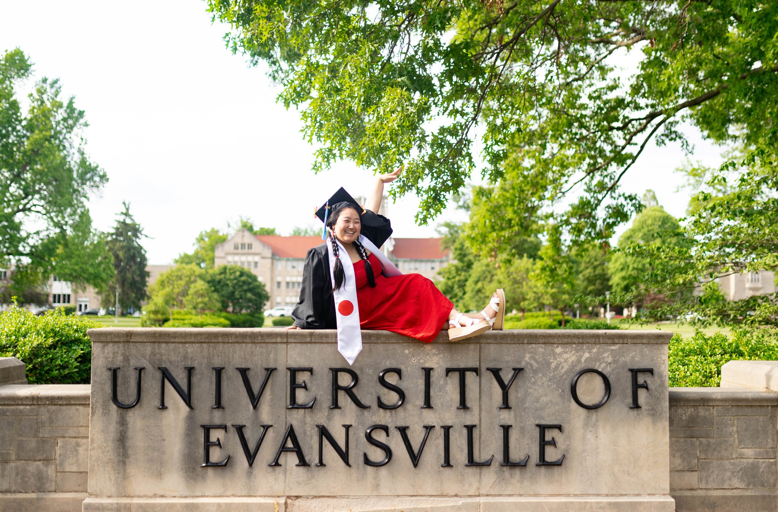 A smiling female graduate in cap and gown sitting on a stone sign that reads 'University of Evansville' outdoors with green trees and university buildings in the background.