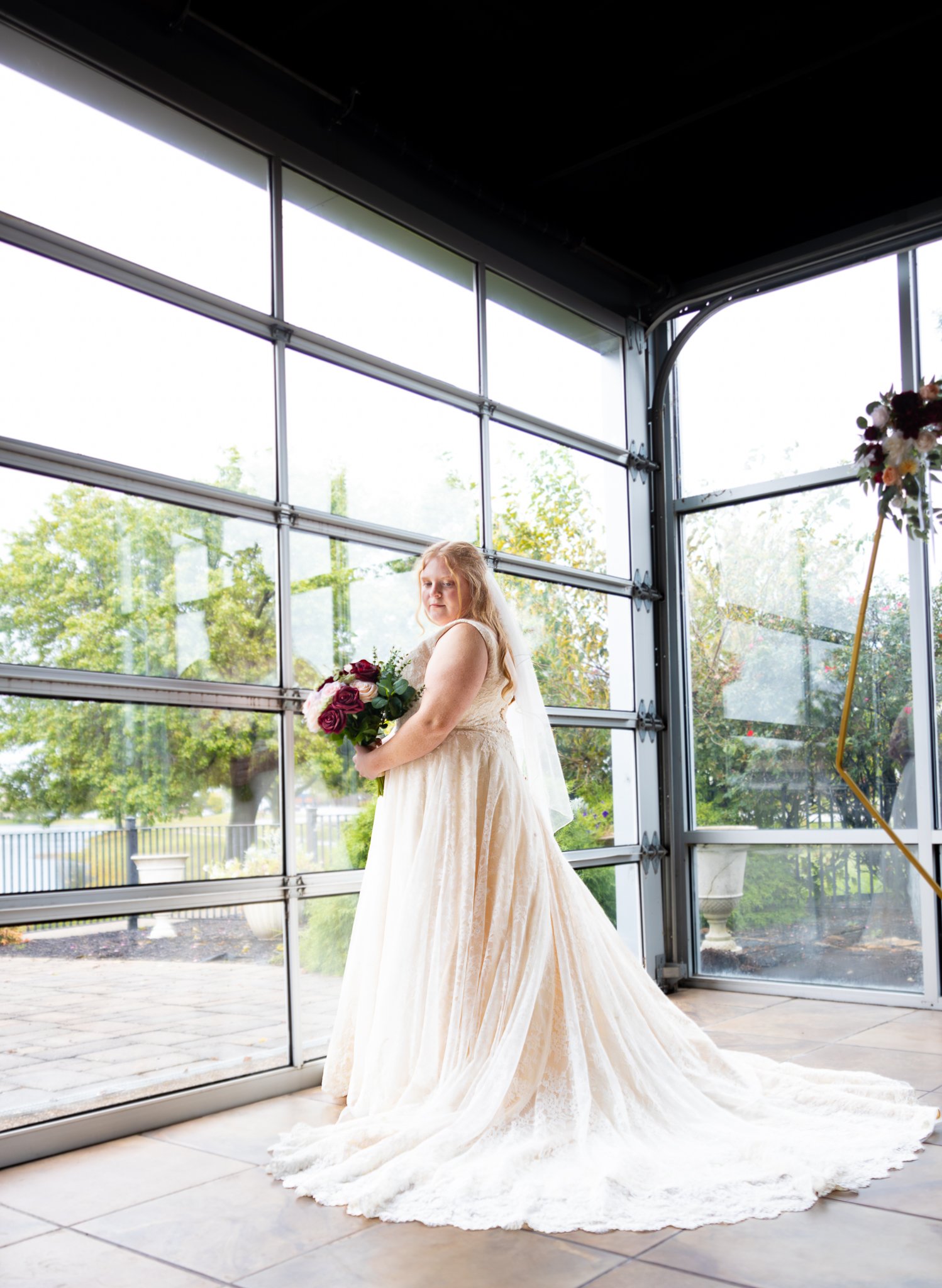 Bride in an ivory wedding dress holding a bouquet, standing inside a modern glass-walled venue with outdoor greenery visible through the windows.